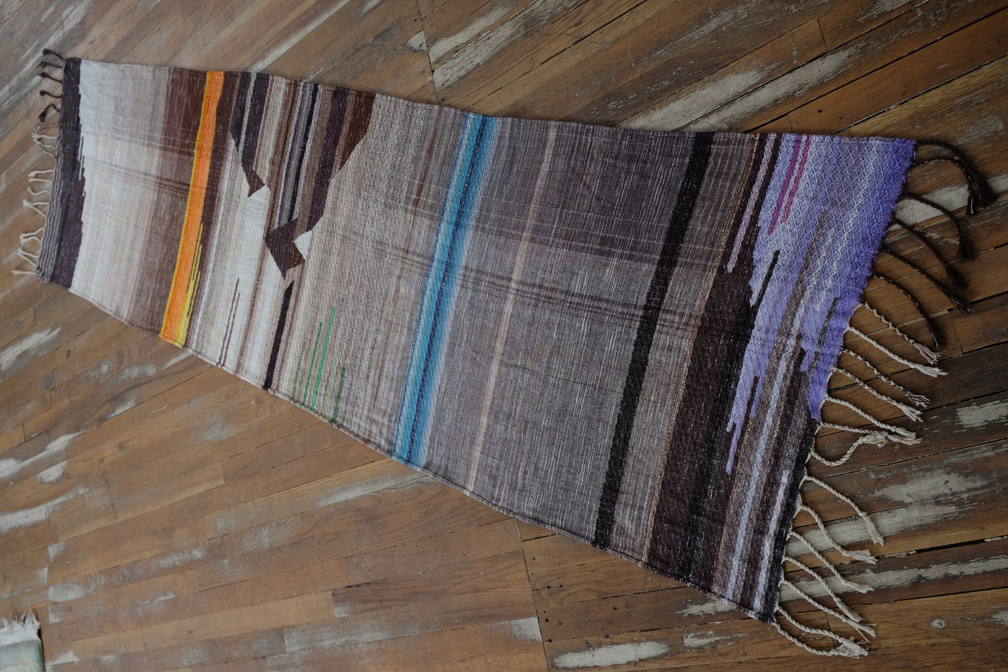 A detail of the geometric tapestry woven designs on a shawl in grayscale, earth-tones and elemental colors of lavender, orange poppy, water and grass. The shawl is laying on a wooden floor