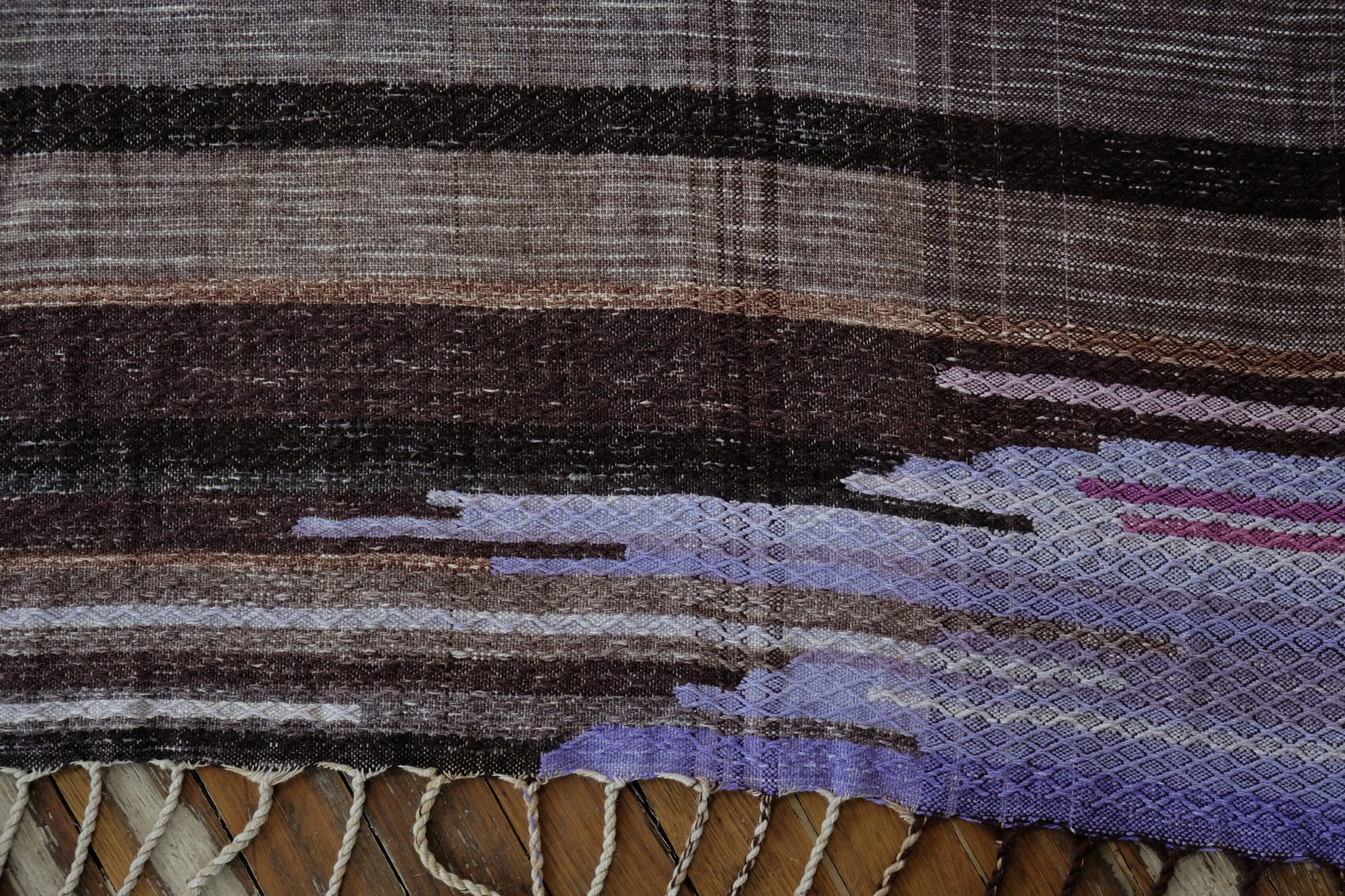 A detail of the geometric tapestry woven designs on a shawl in grayscale, earth-tones and elemental colors of lavender, orange poppy, water and grass. The shawl is laying on a wooden floor