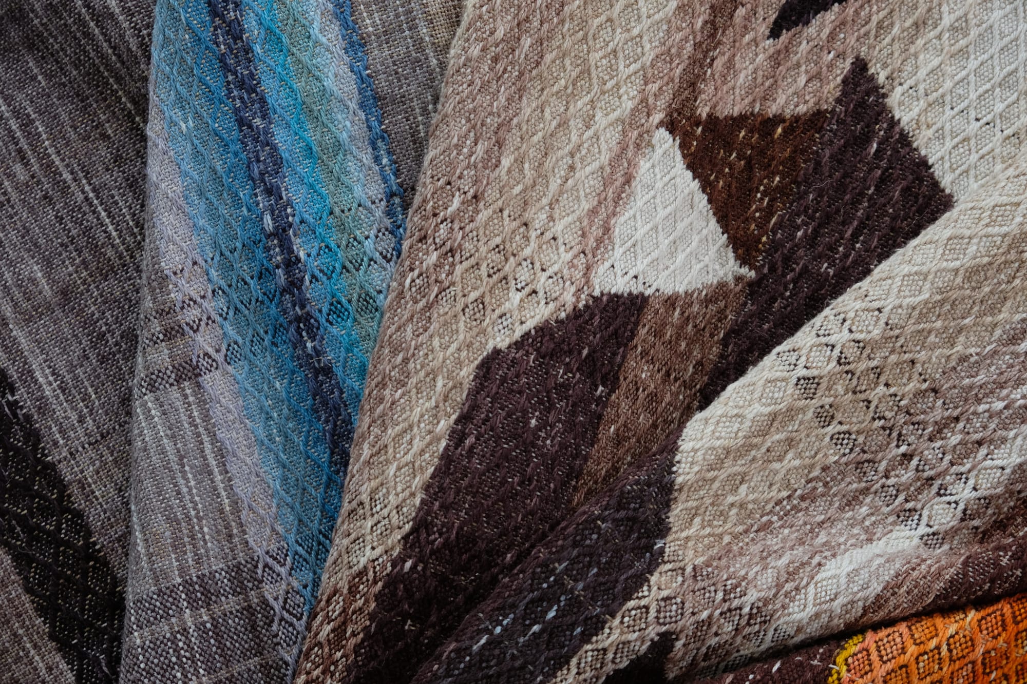 A detail of the geometric tapestry woven designs on a shawl in grayscale, earth-tones and elemental colors of lavender, orange poppy, water and grass. The shawl is laying on a wooden floor