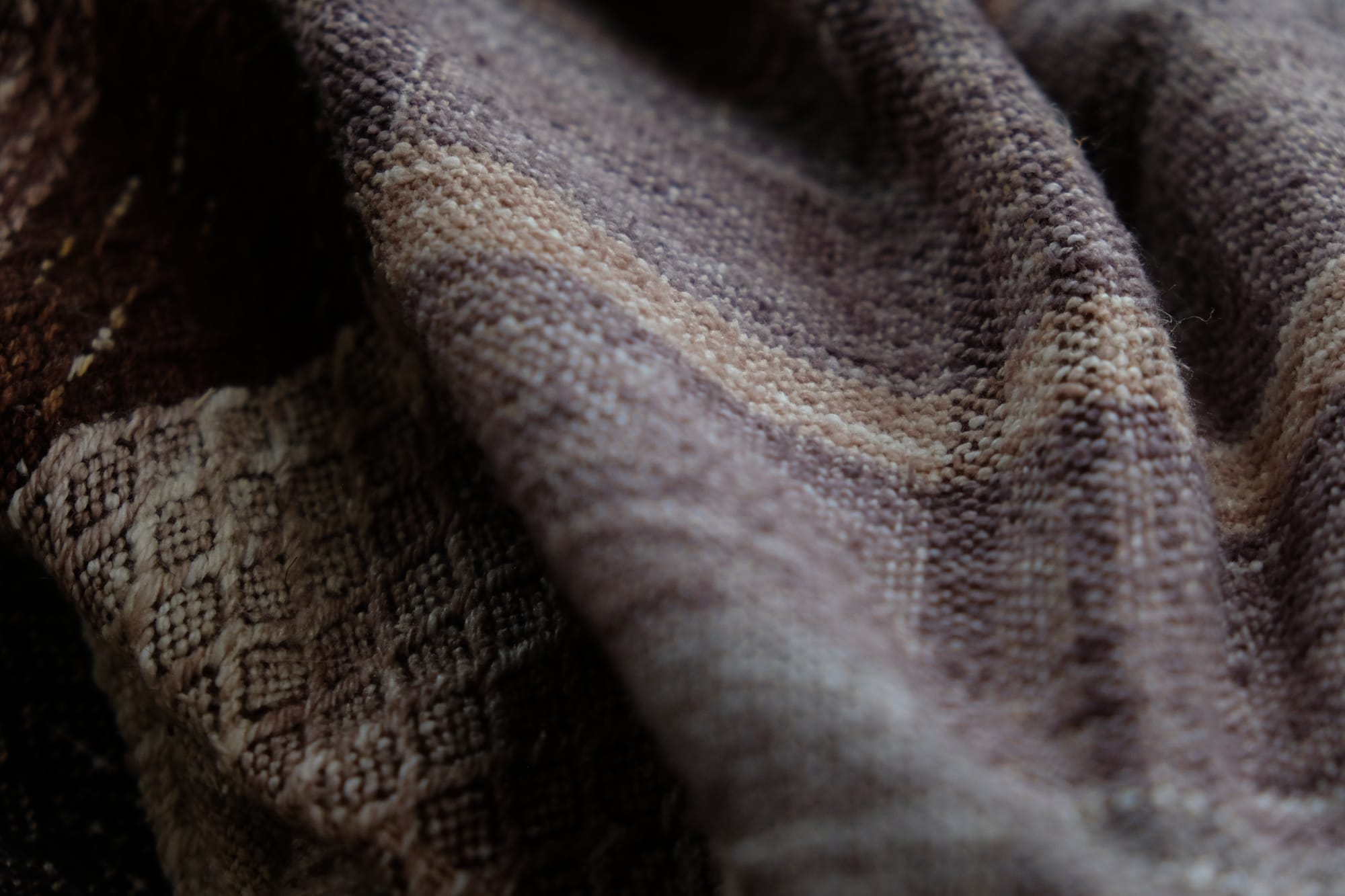 A detail of the geometric tapestry woven designs on a shawl in grayscale, earth-tones and elemental colors of lavender, orange poppy, water and grass. The shawl is laying on a wooden floor