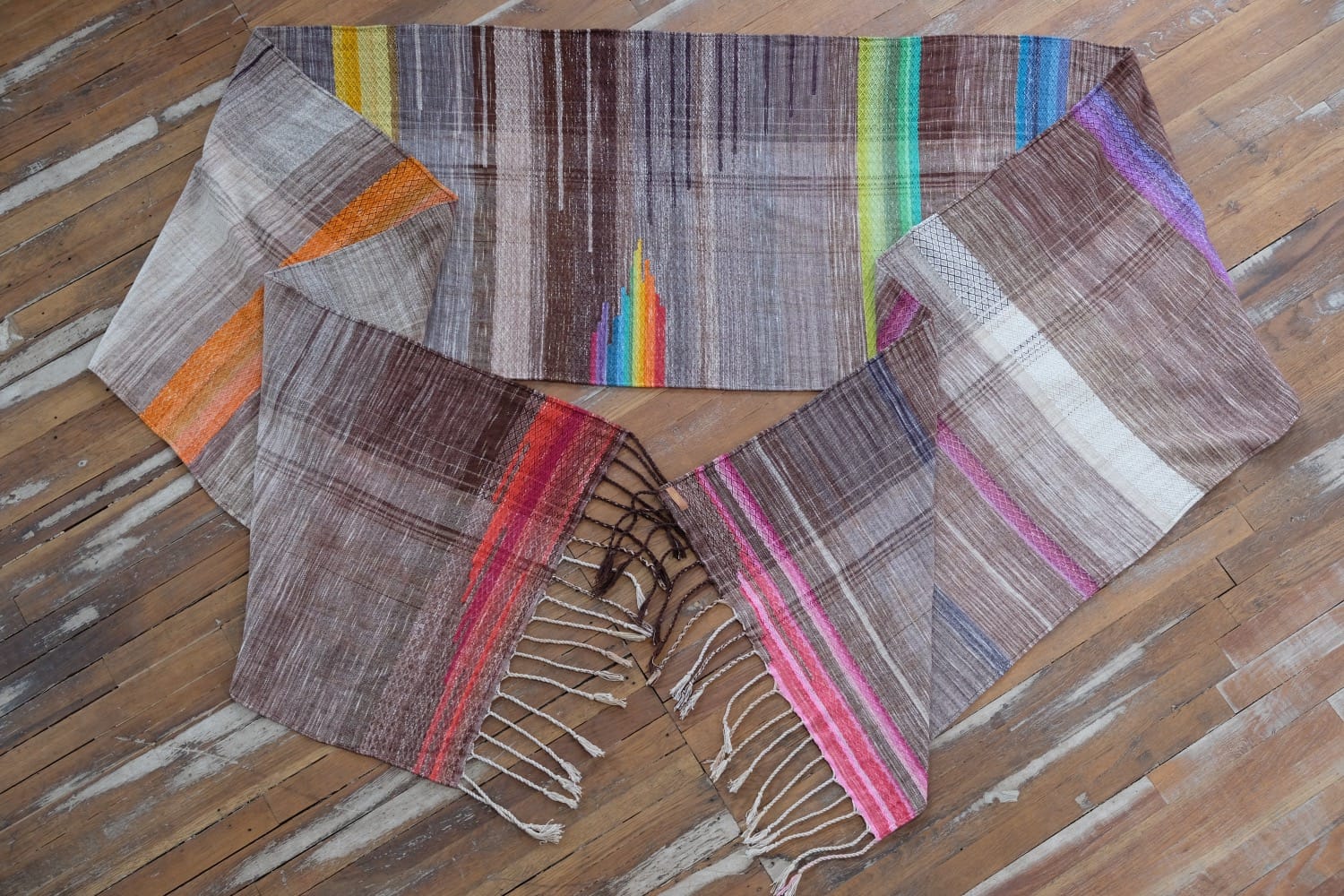 A detail of a length of handwoven fabric in earth-tones and all the colors of the rainbow in prismatic sequence, laying on a wood floor