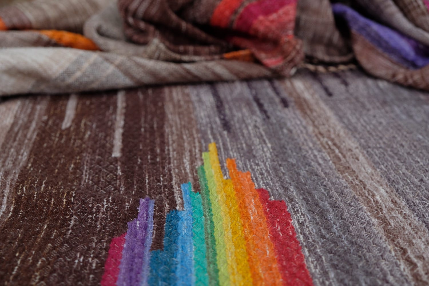 A detail of a length of handwoven fabric in earth-tones and all the colors of the rainbow in prismatic sequence, laid out on a wood floor