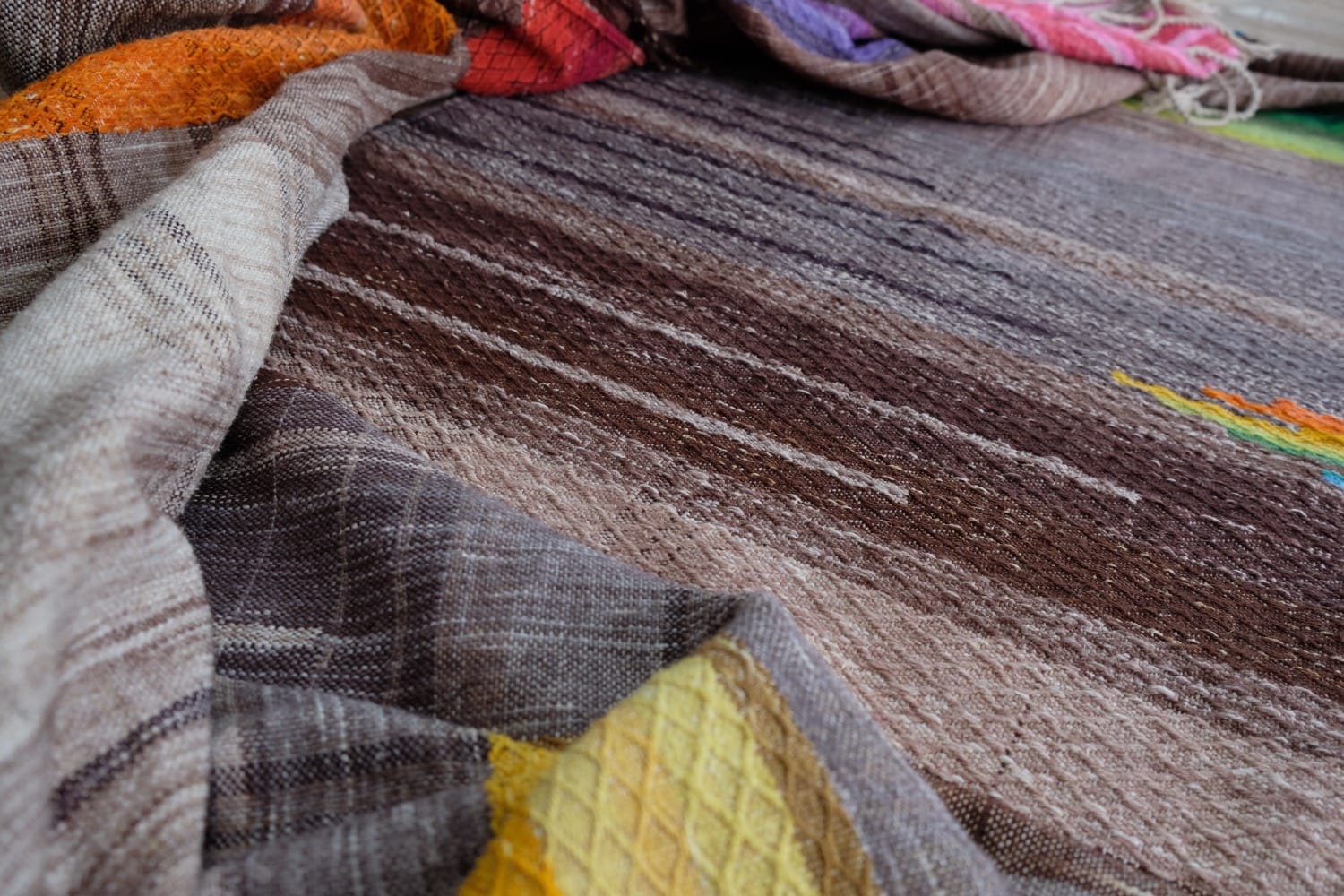 A detail of a length of handwoven fabric in earth-tones and all the colors of the rainbow in prismatic sequence, laid out on a wood floor