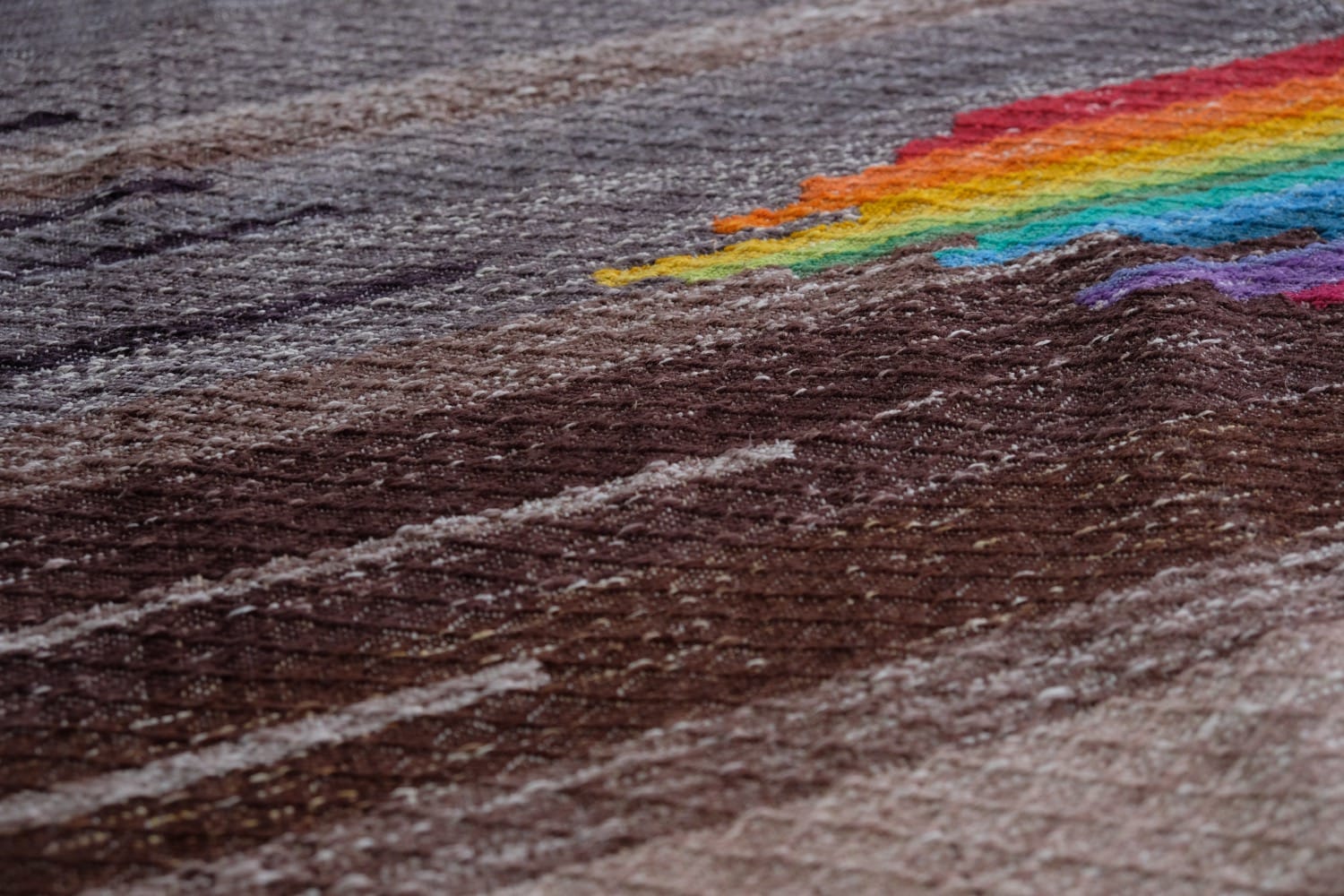 A detail of a length of handwoven fabric in earth-tones and all the colors of the rainbow in prismatic sequence, laid out on a wood floor