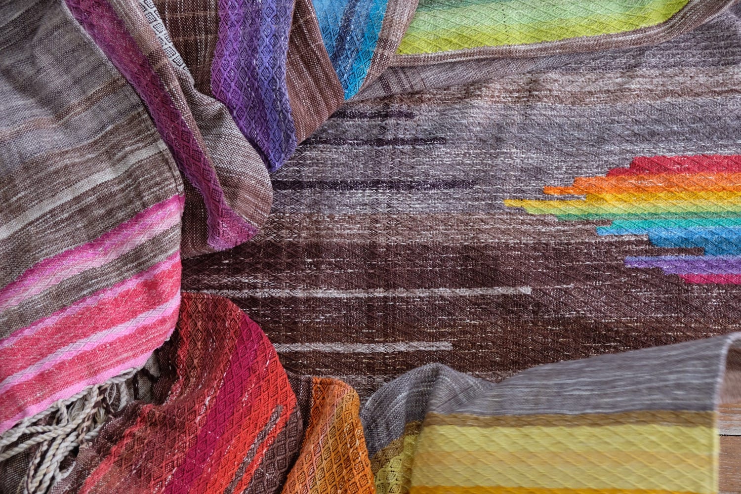 A detail of a length of handwoven fabric in earth-tones and all the colors of the rainbow in prismatic sequence, laid out on a wood floor