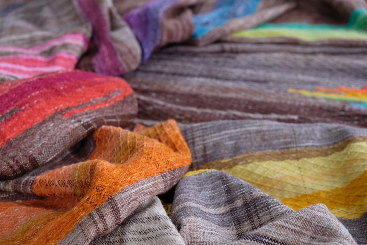 A detail of a length of handwoven fabric in earth-tones and all the colors of the rainbow in prismatic sequence, laid out on a wood floor