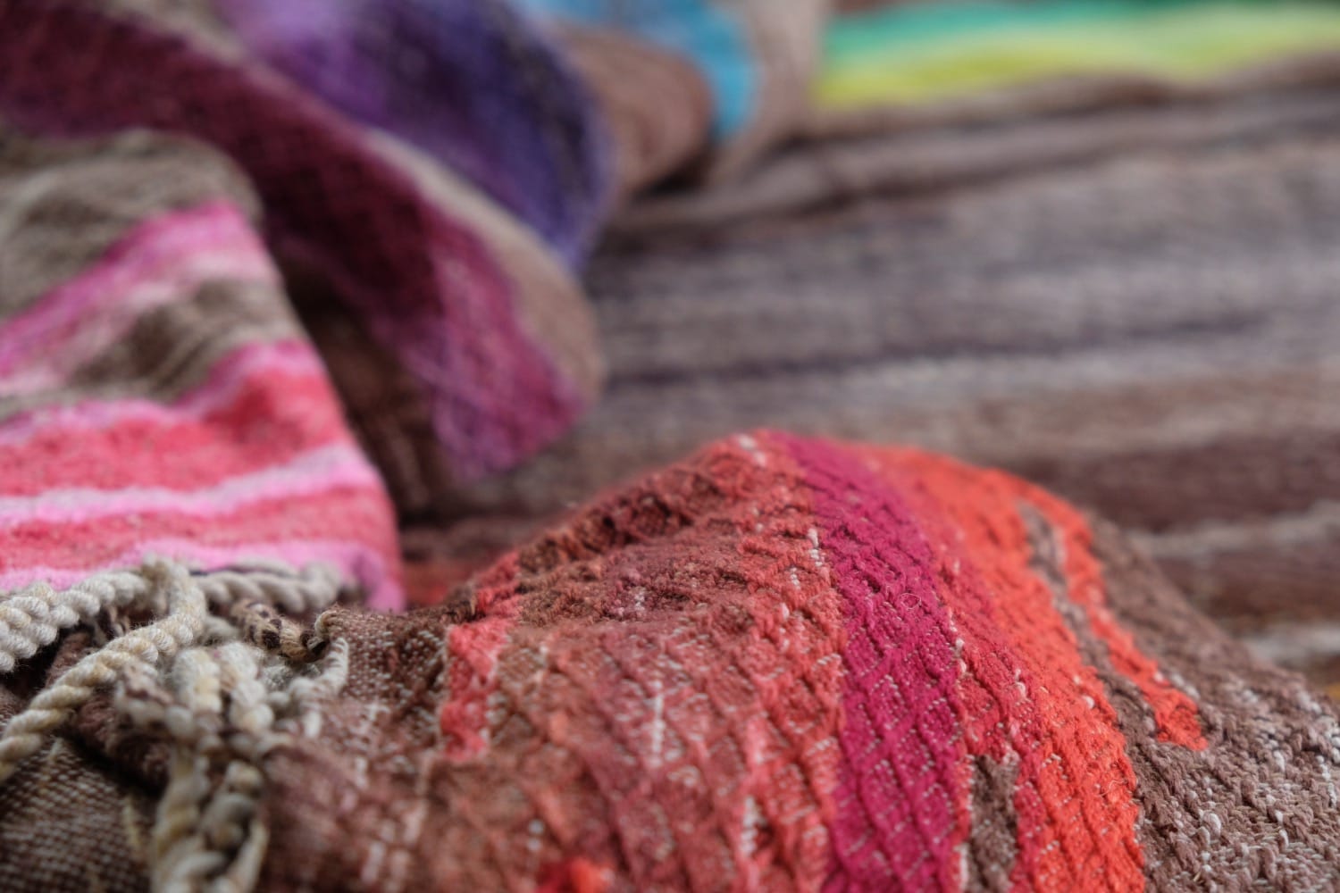 A detail of a length of handwoven fabric in earth-tones and all the colors of the rainbow in prismatic sequence, laid out on a wood floor