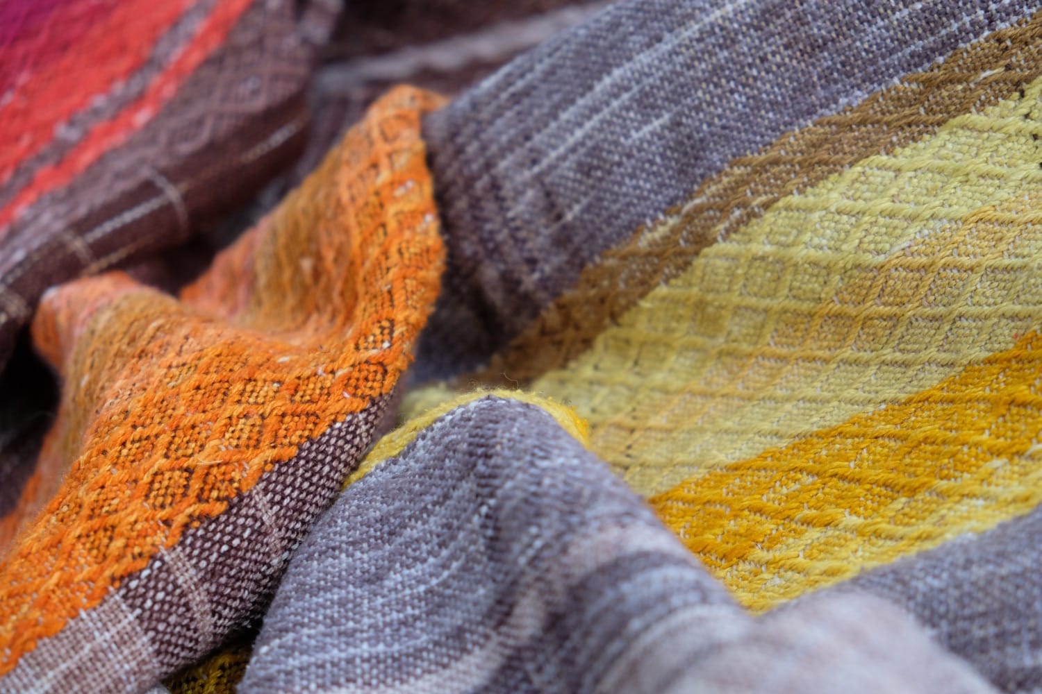 A detail of a length of handwoven fabric in earth-tones and all the colors of the rainbow in prismatic sequence, laid out on a wood floor