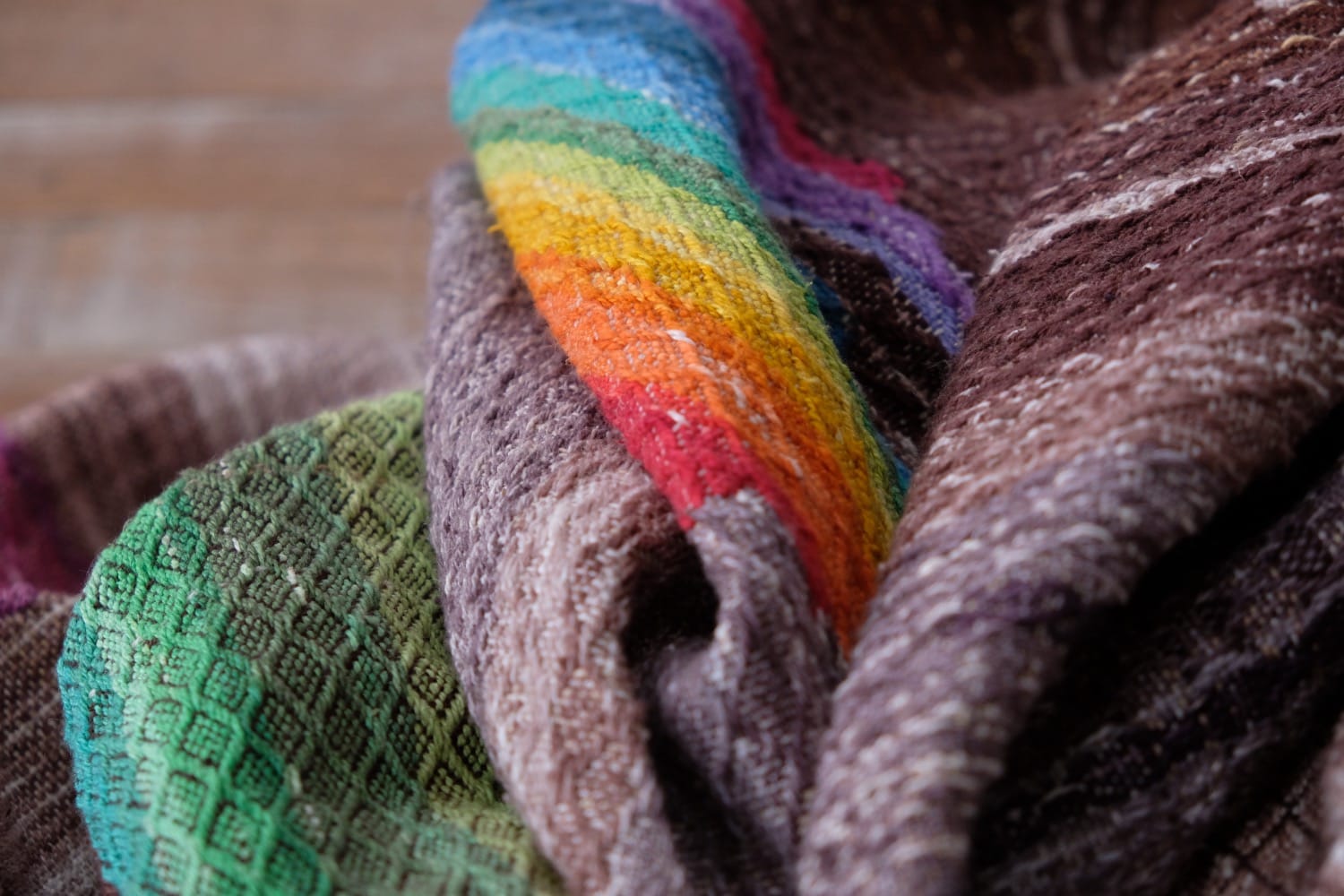 A detail of a length of handwoven fabric in earth-tones and all the colors of the rainbow in prismatic sequence, laid out on a wood floor