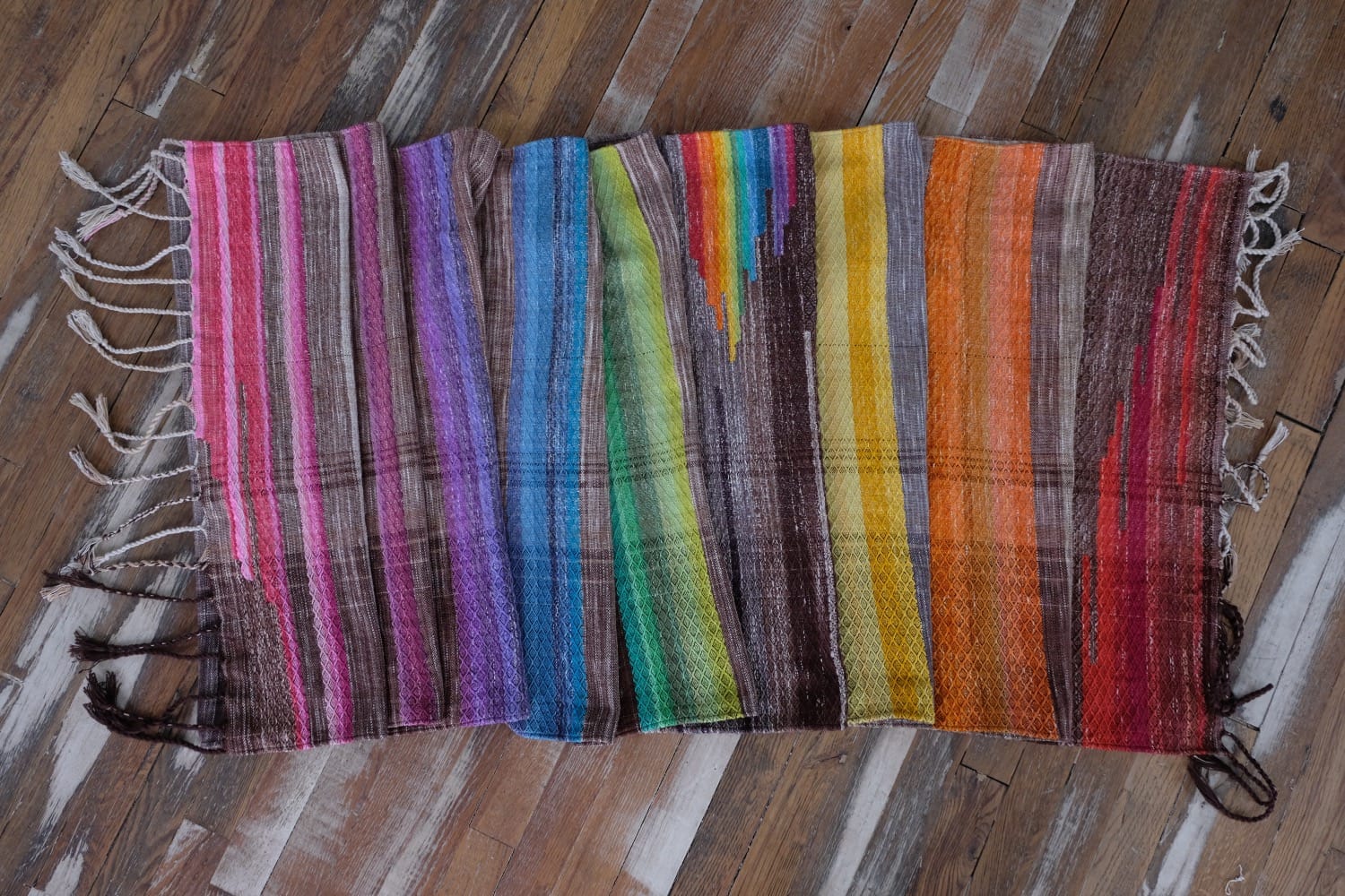 A detail of a length of handwoven fabric in earth-tones and all the colors of the rainbow in prismatic sequence, laying on a wood floor