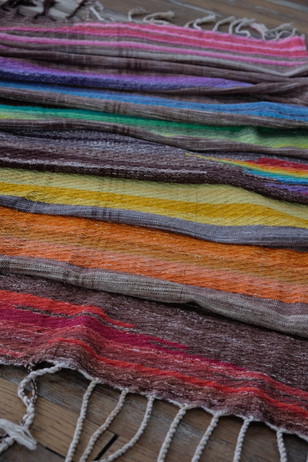 A detail of a length of handwoven fabric in earth-tones and all the colors of the rainbow in prismatic sequence, laying on a wood floor