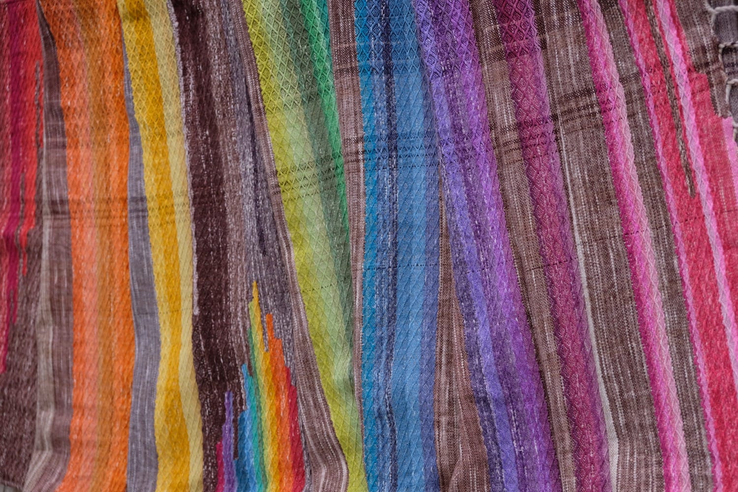 A detail of a length of handwoven fabric in earth-tones and all the colors of the rainbow in prismatic sequence, laid out on a wood floor