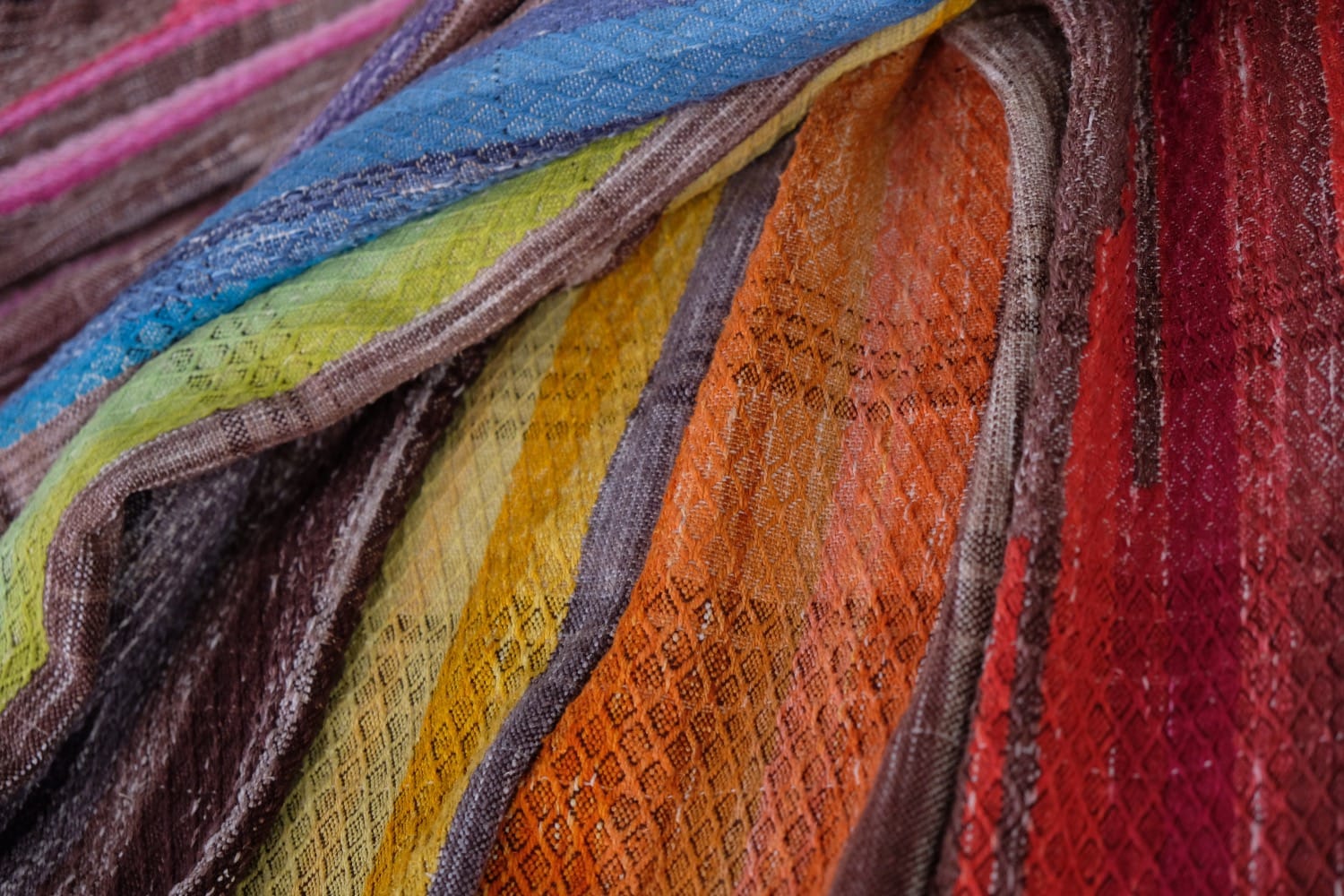 A detail of a length of handwoven fabric in earth-tones and all the colors of the rainbow in prismatic sequence, laid out on a wood floor