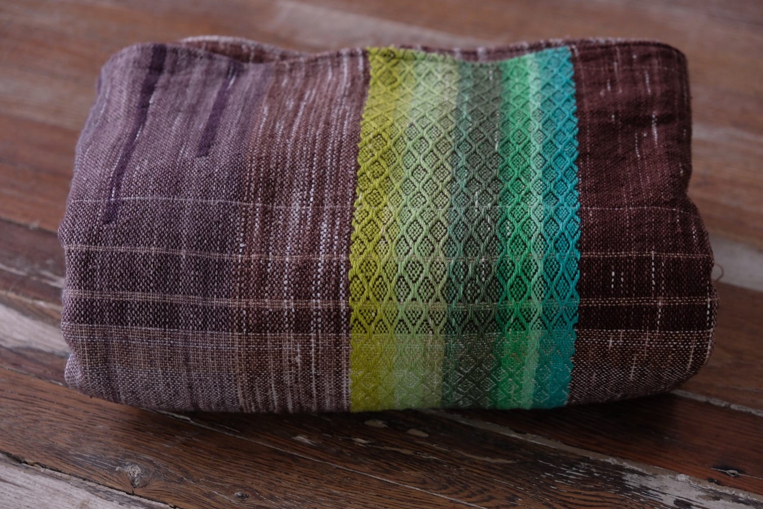 A detail of a length of handwoven fabric in earth-tones and all the colors of the rainbow in prismatic sequence, folded on a wood floor