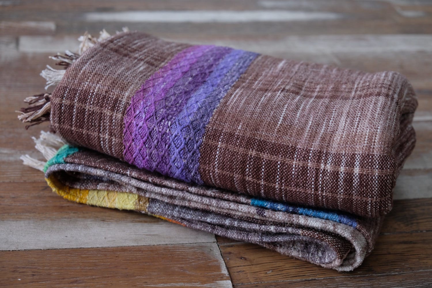 A detail of a length of handwoven fabric in earth-tones and all the colors of the rainbow in prismatic sequence, folded on a wood floor