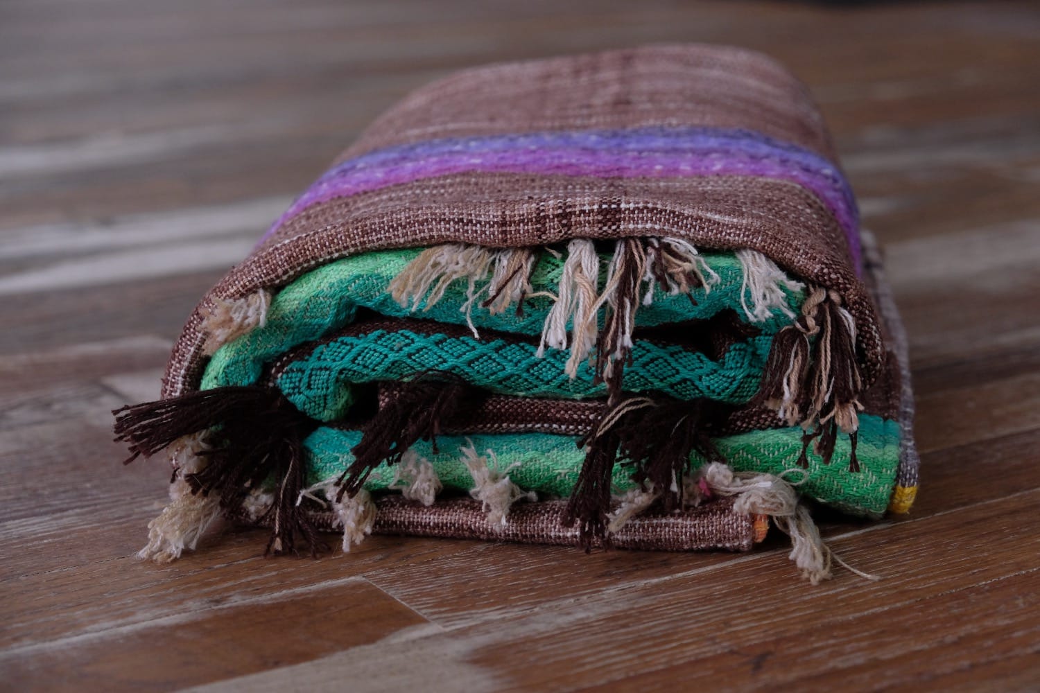 A detail of a length of handwoven fabric in earth-tones and all the colors of the rainbow in prismatic sequence, folded on a wood floor