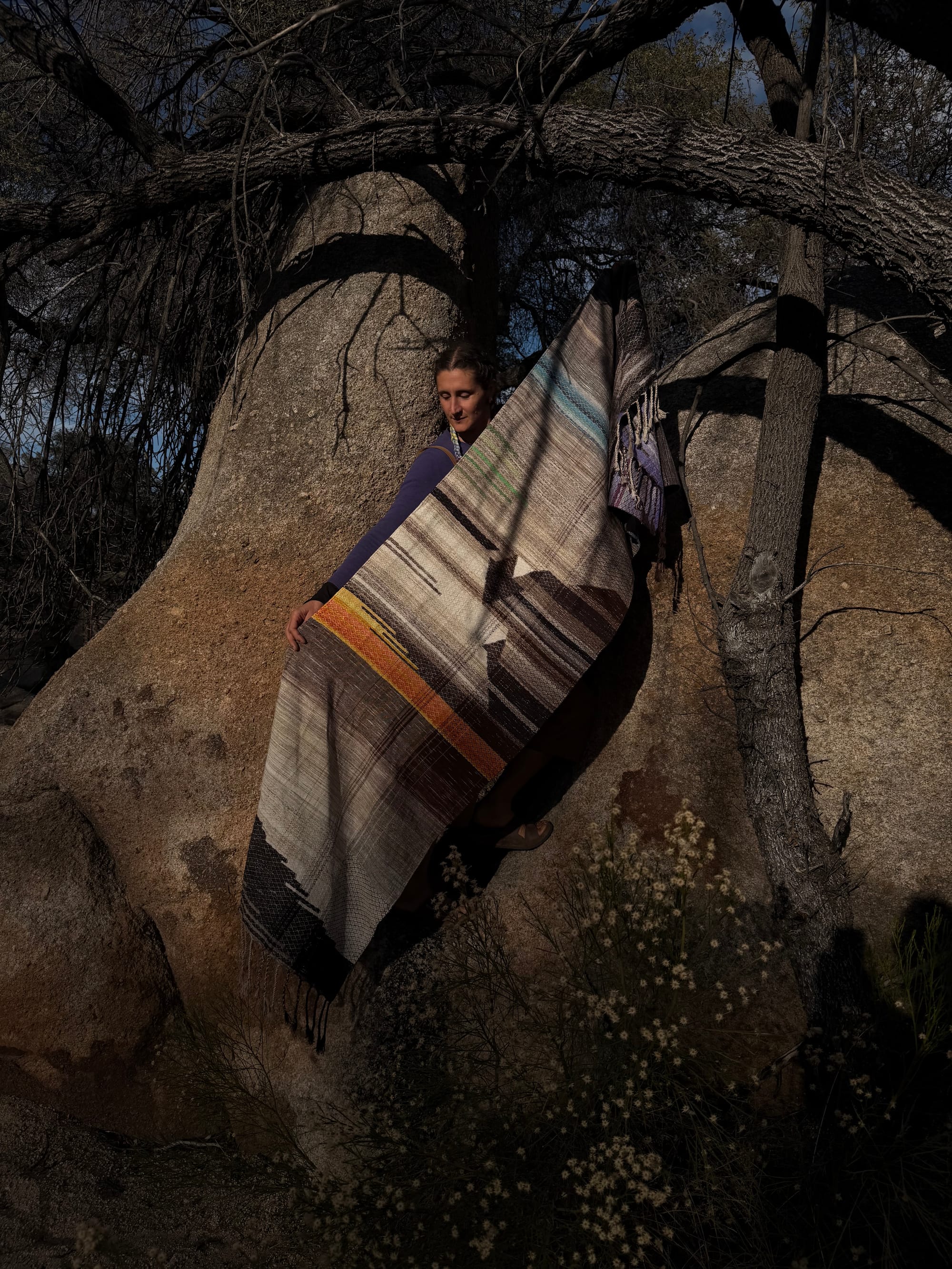 A woman stands in the hollow of a giant granite boulder wearing a handwoven shawl in tones of grey, brown, blue water, orange poppies, lavender flowers and sprigs of grass