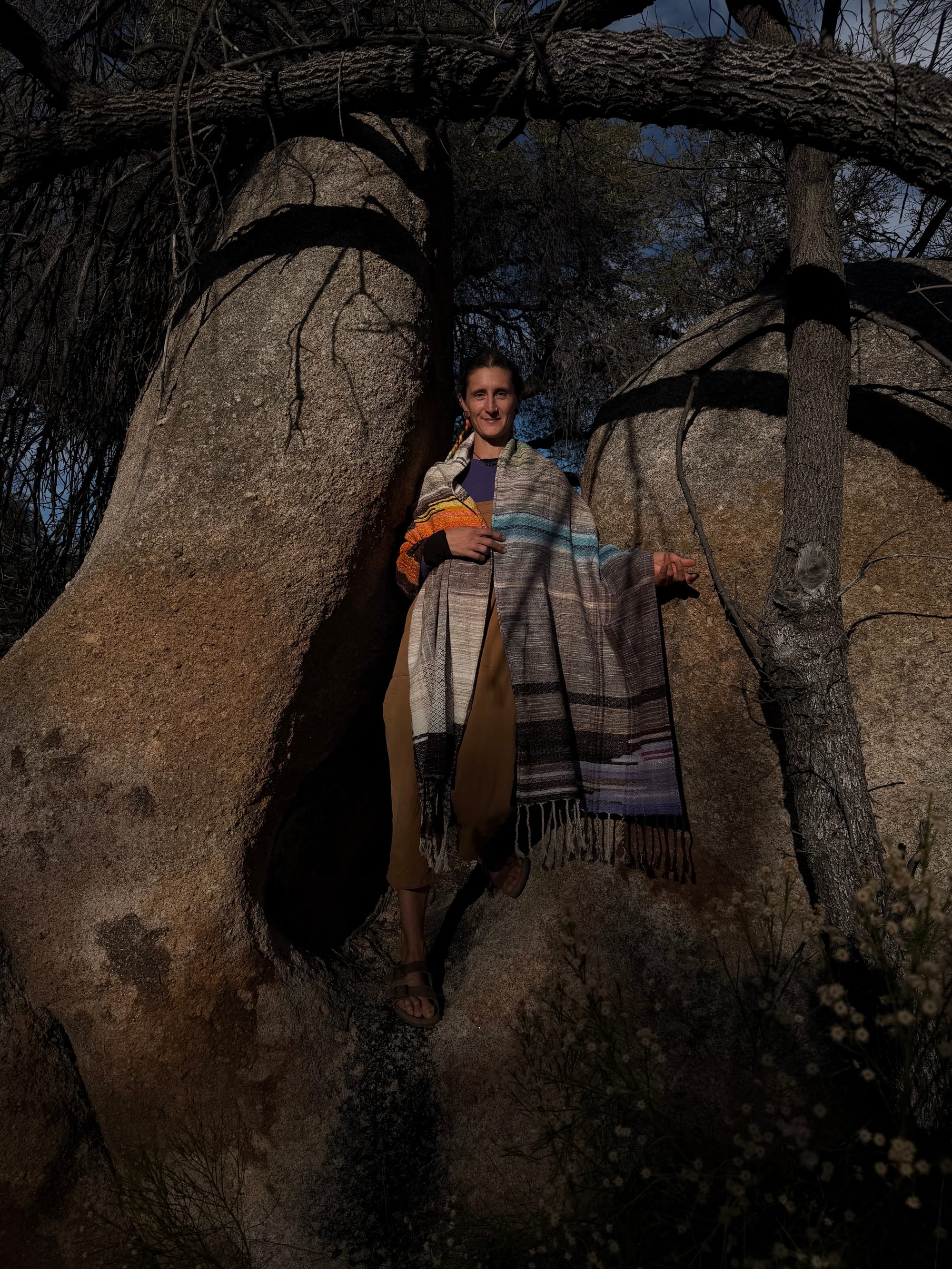 A woman stands in the hollow of a giant granite boulder wearing a handwoven shawl in tones of grey, brown, blue water, orange poppies, lavender flowers and sprigs of grass