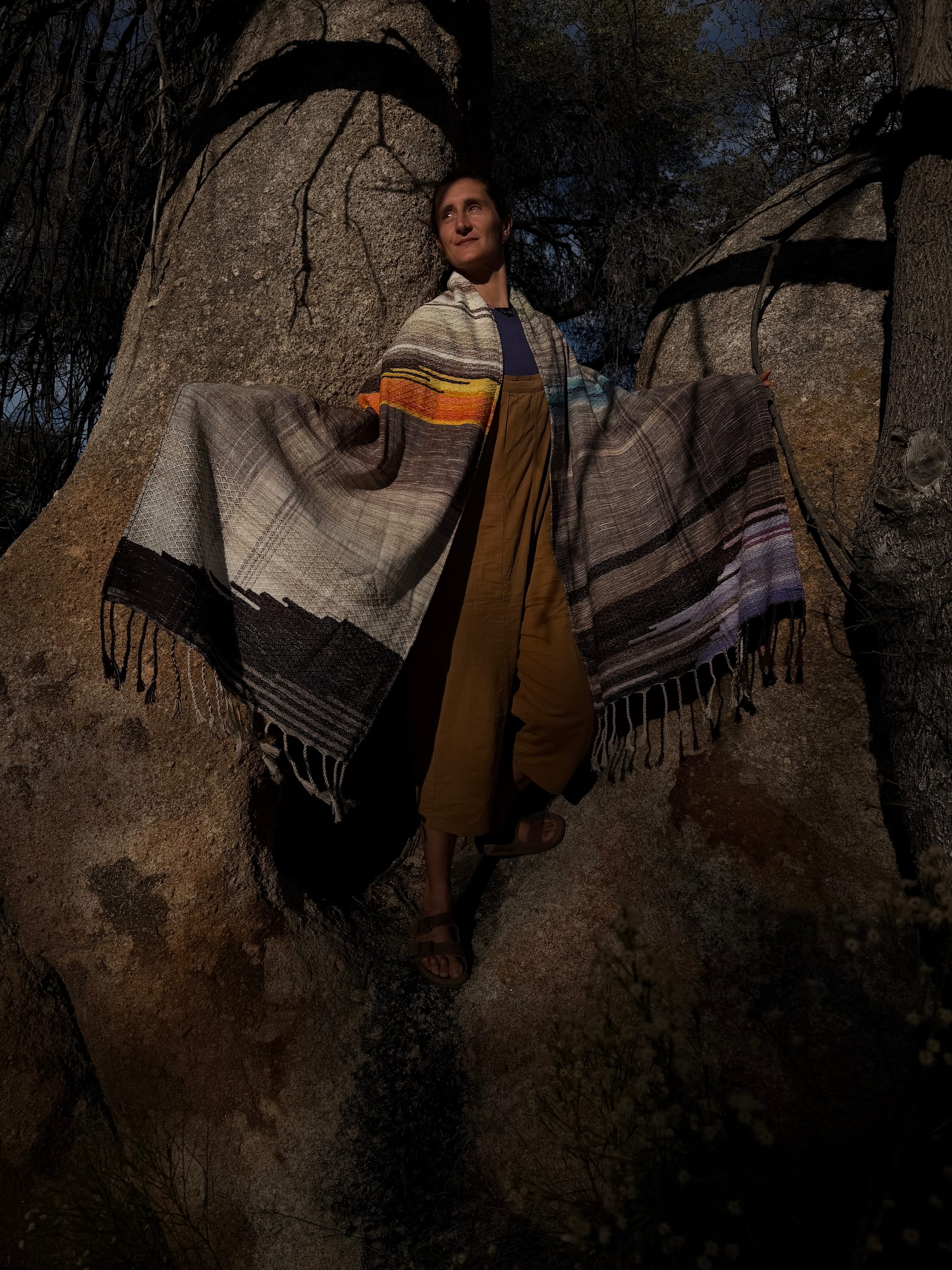 A woman stands in the hollow of a giant granite boulder wearing a handwoven shawl in tones of grey, brown, blue water, orange poppies, lavender flowers and sprigs of grass