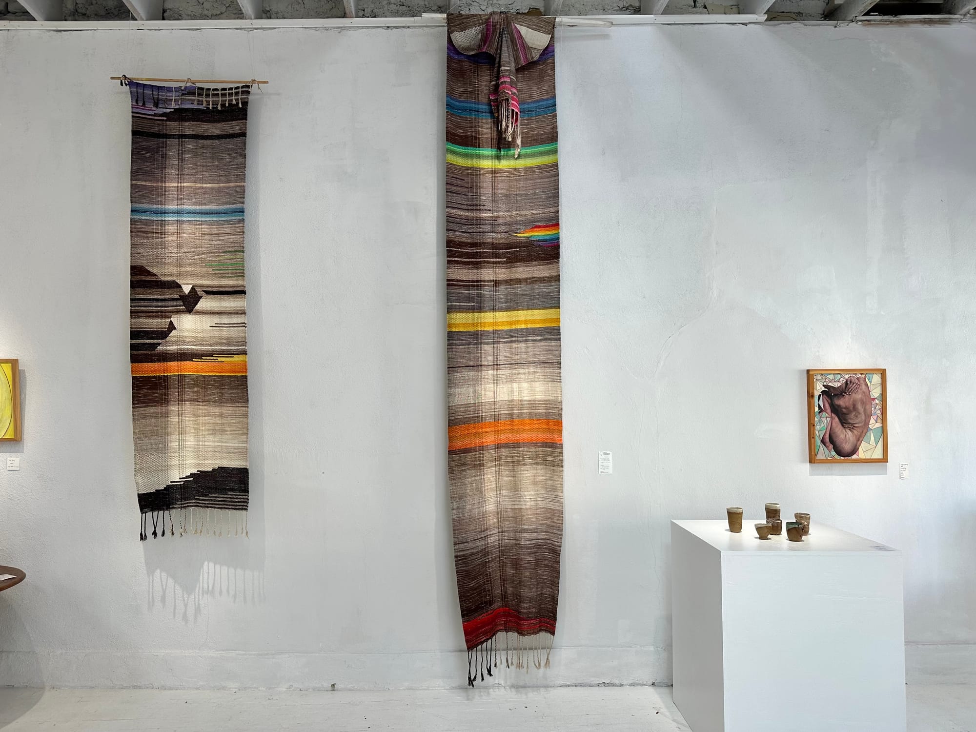Two handwoven multi-colored tapestries hang on the wall of a white gallery space. There are also two paintings on the walls, and marble sculptures on pedestals