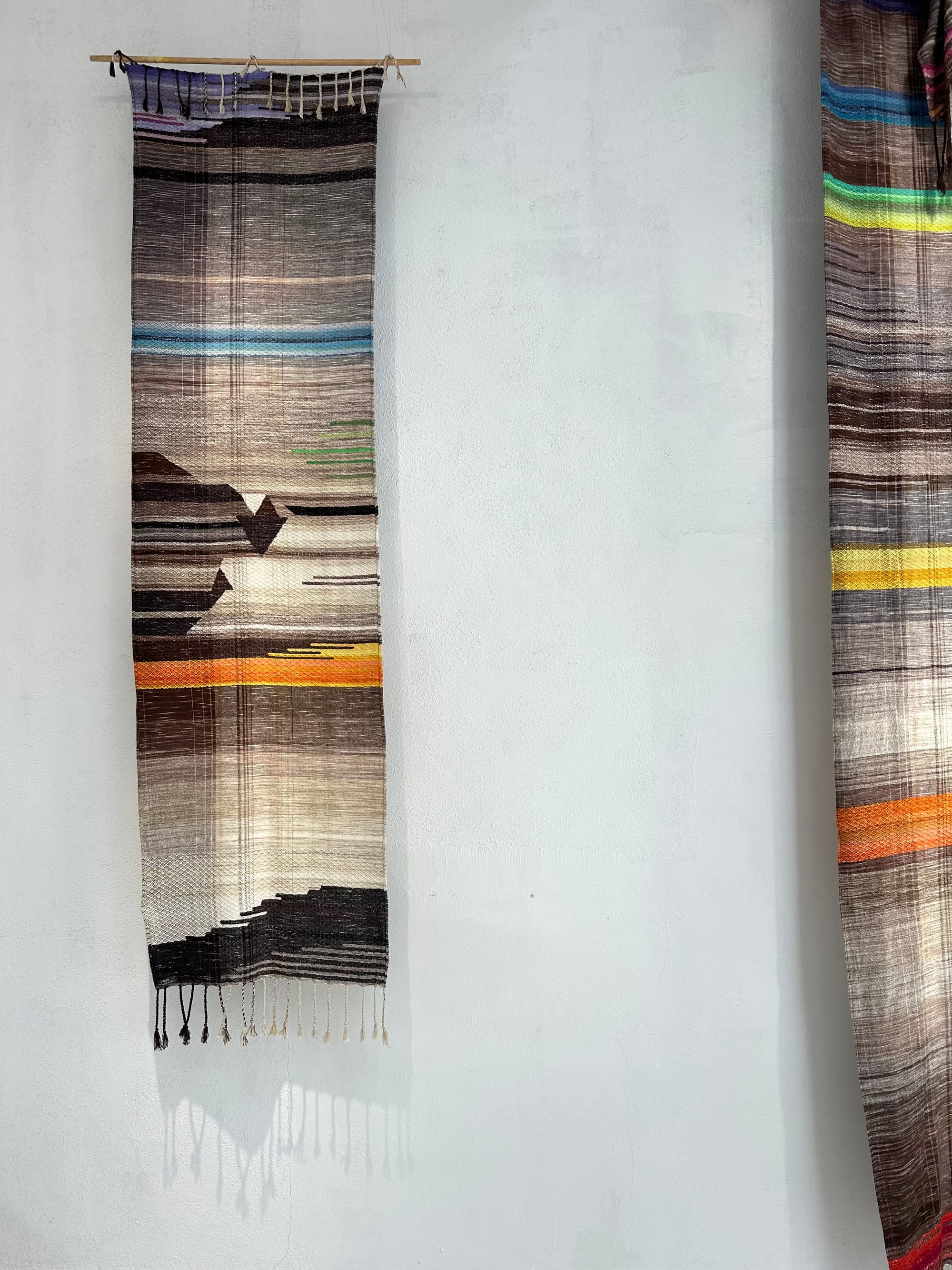 two handwoven lengths of fabric hang on a gallery wall, the weavings are earth-toned with pops of the colors of the rainbow.