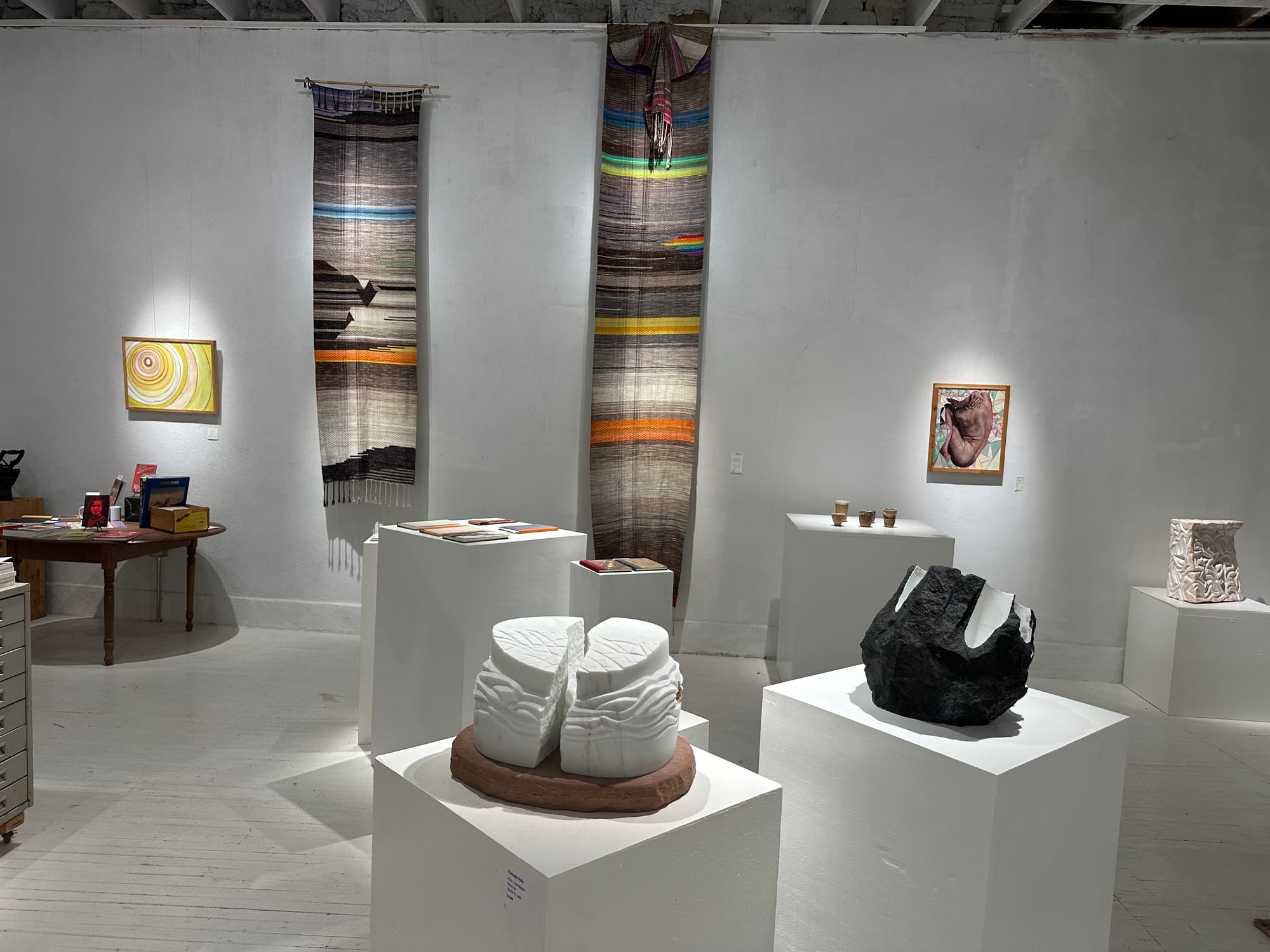 Two handwoven multi-colored tapestries hang on the wall of a white gallery space. There are also two paintings on the walls, and marble sculptures on pedestals