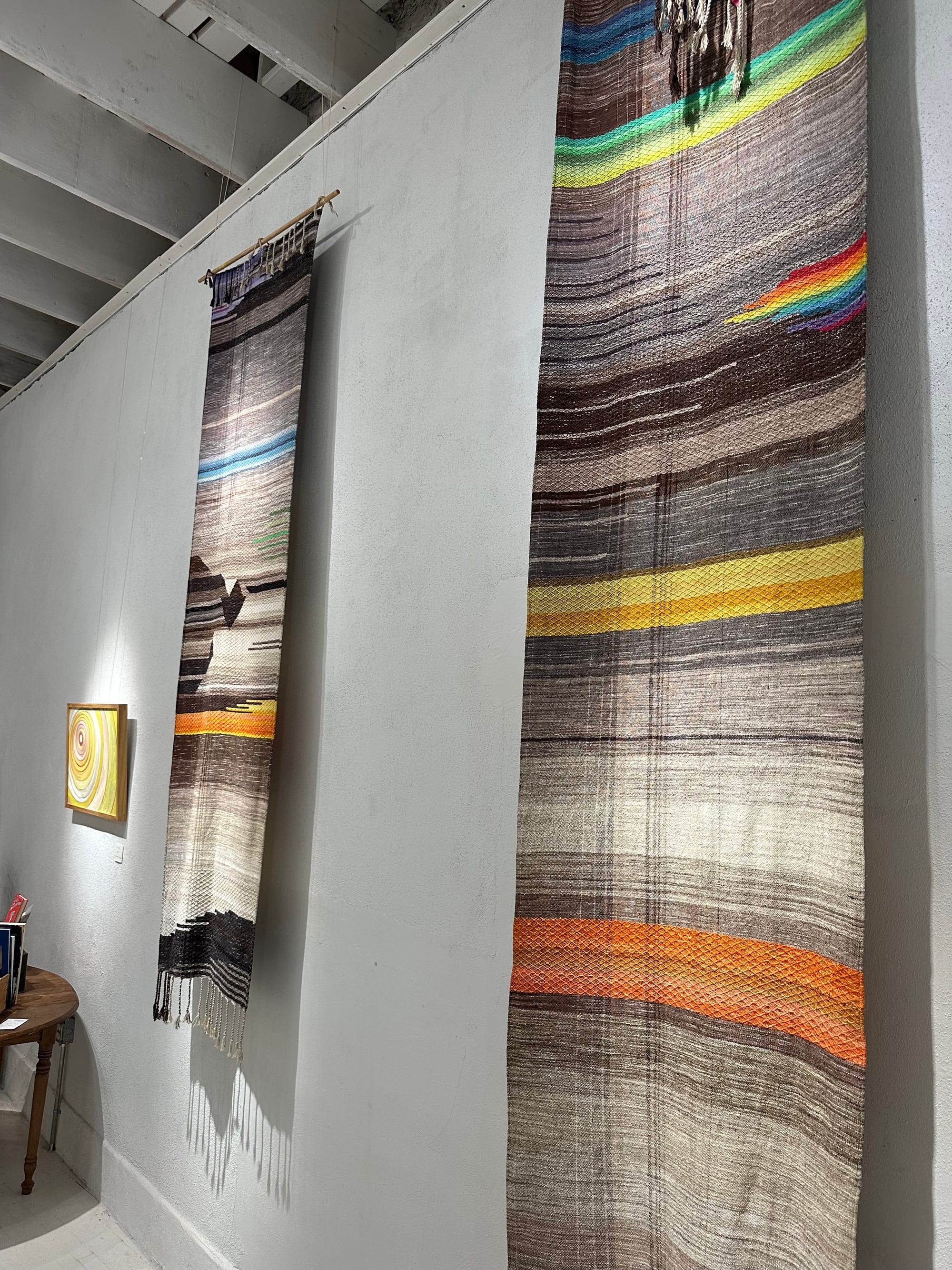 Two handwoven multi-colored tapestries hang on the wall of a white gallery space. There are also two paintings on the walls, and marble sculptures on pedestals