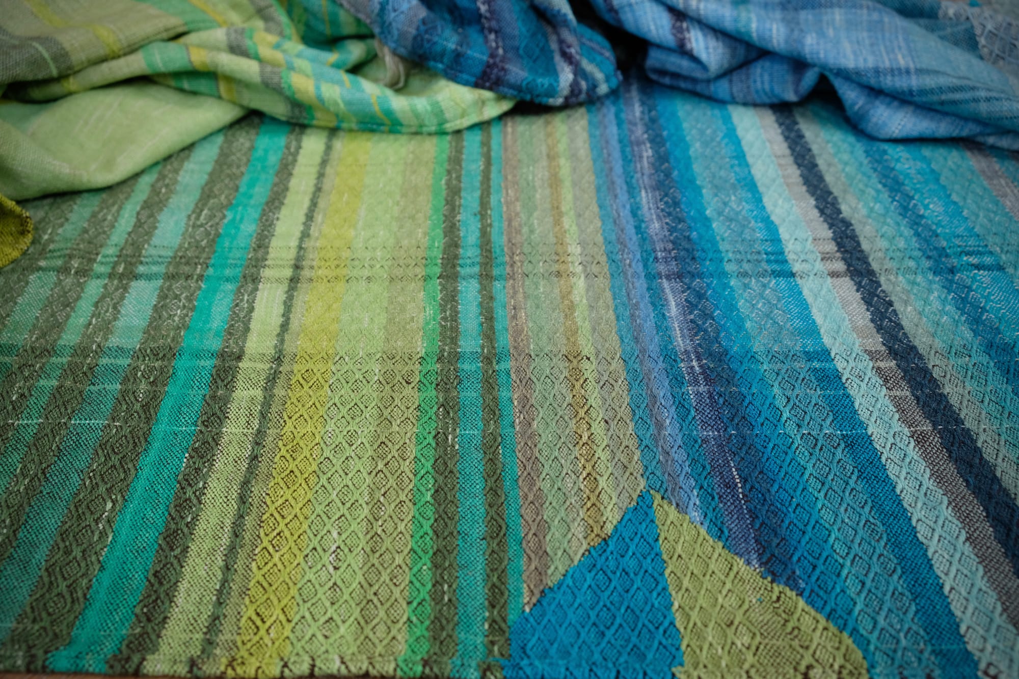 a detail of the pyramid tapestry woven detail in the middle of on an oak wood floor is laid 5 meters of handwoven fabric in textured stripes of greens on one side and shades of blue on the other