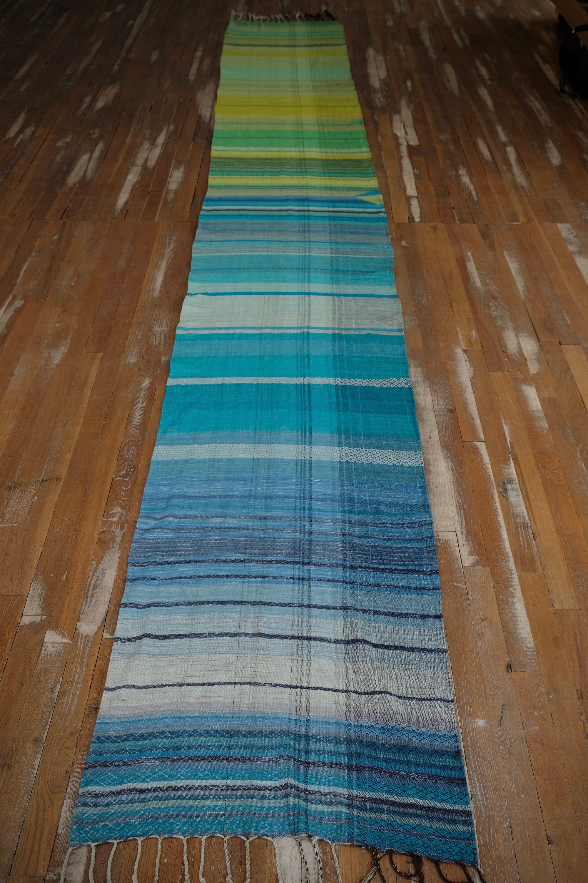 on an oak wood floor is laid 5 meters of handwoven fabric in textured stripes of greens on one side and shades of blue on the other