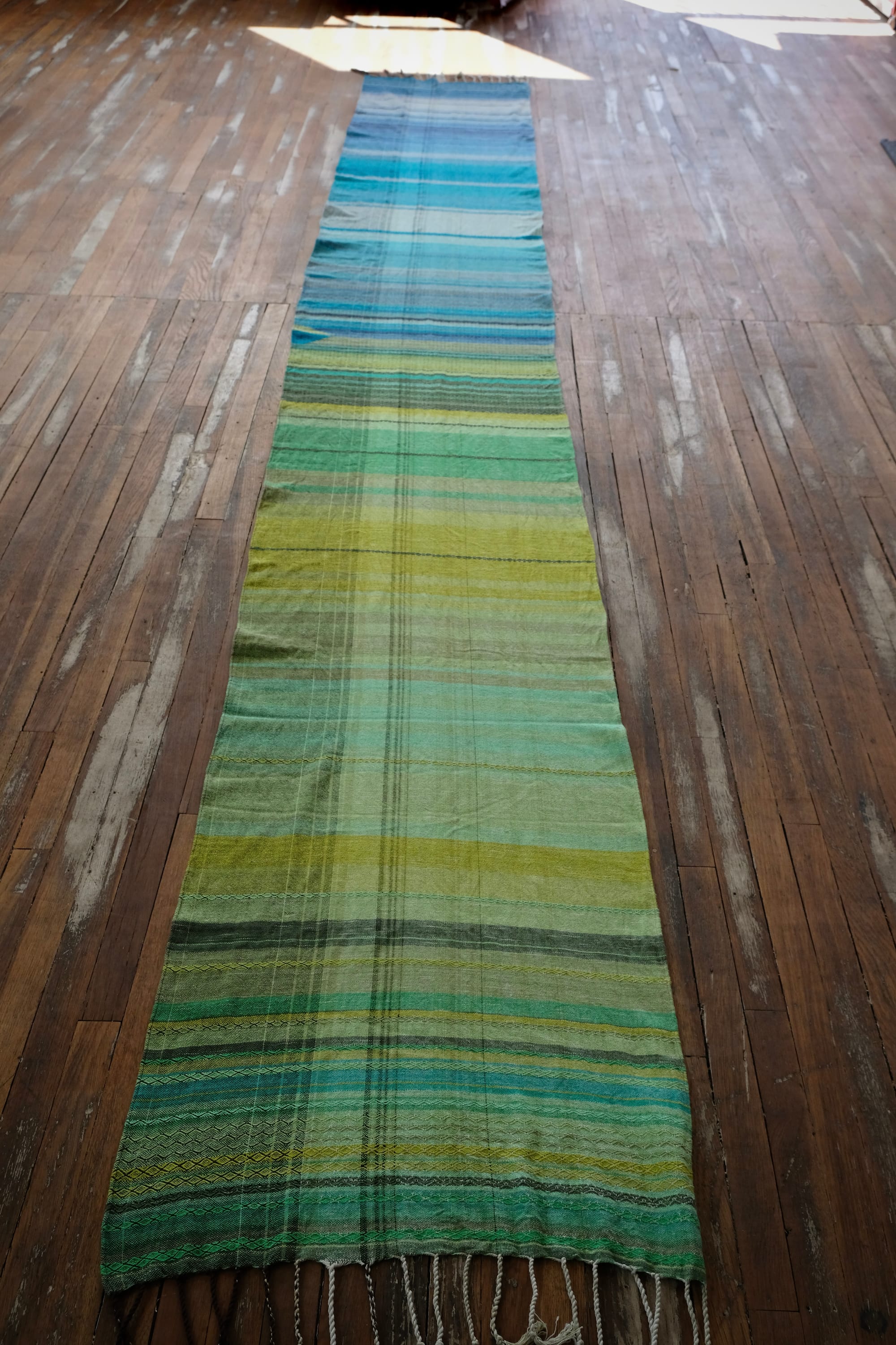 on an oak wood floor is laid 5 meters of handwoven fabric in textured stripes of greens on one side and shades of blue on the other