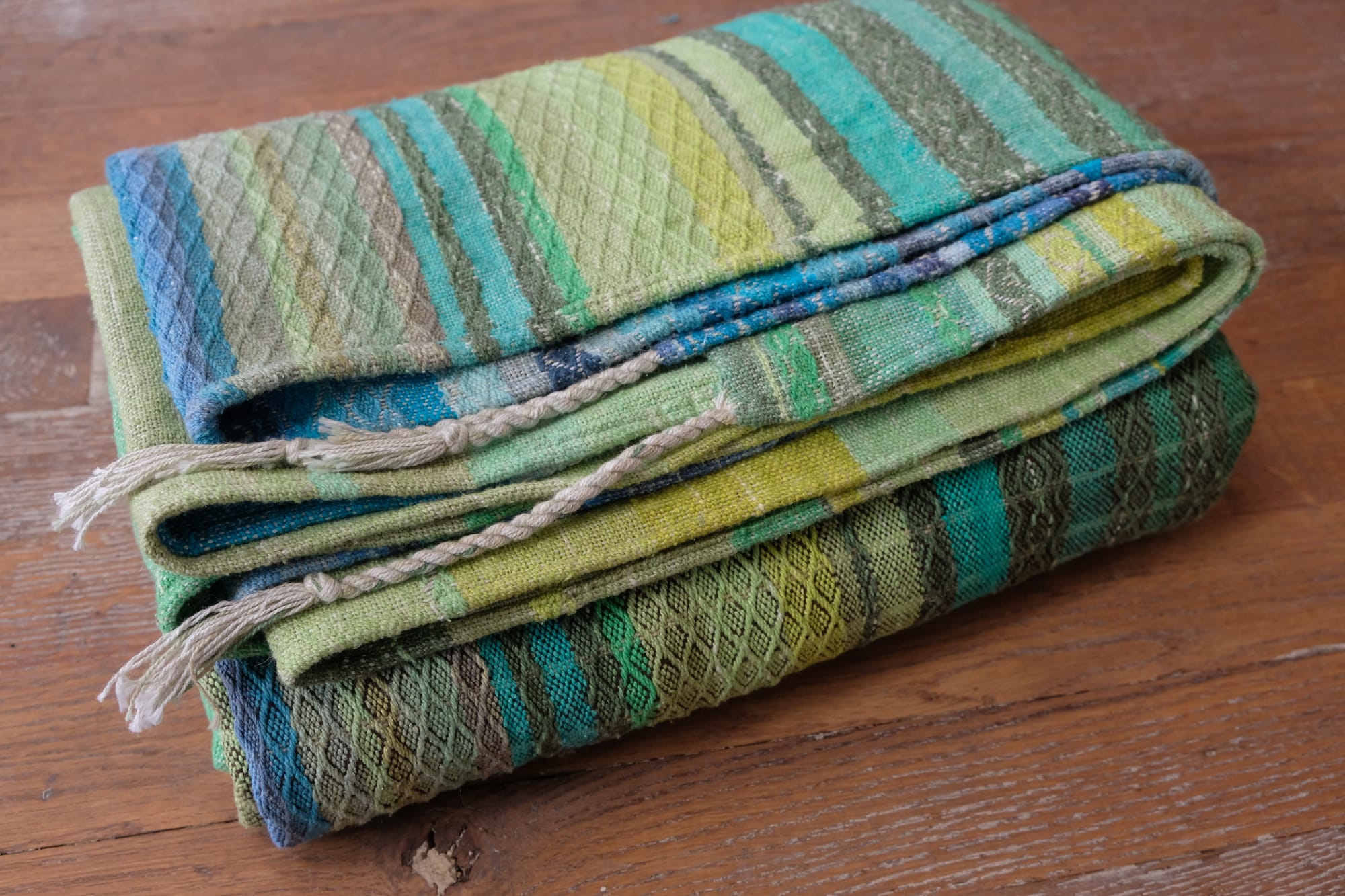 A folded bundle of handwoven fabric rests on an oak wood floor. The part of the fabric you can see is striped shades of blue and green with textured diamond pattern stripes