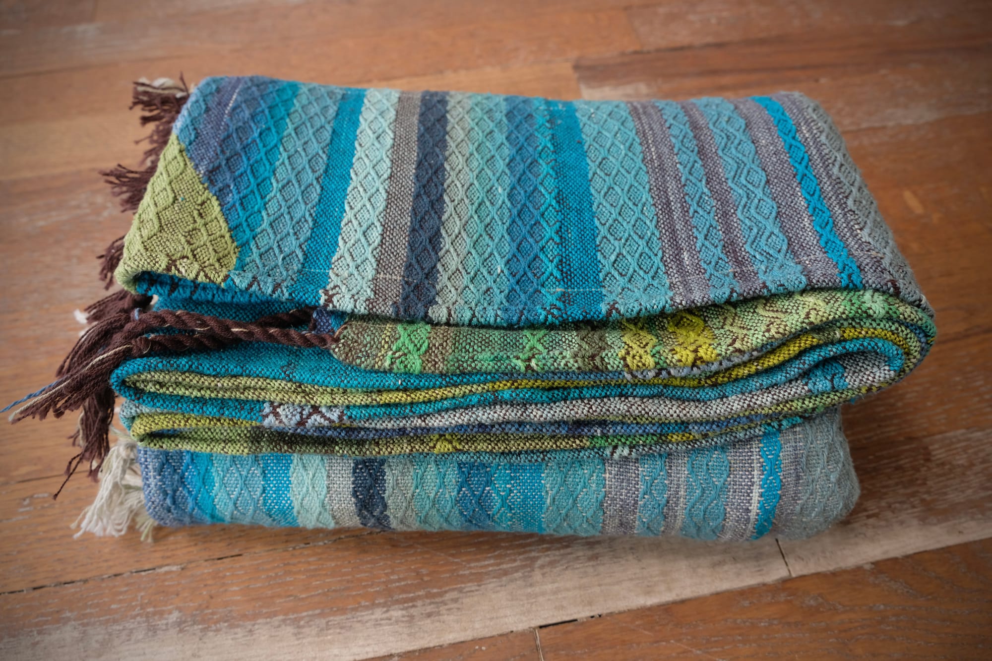 A folded bundle of handwoven fabric rests on an oak wood floor. The part of the fabric you can see is striped shades of blue and green with textured diamond pattern stripes