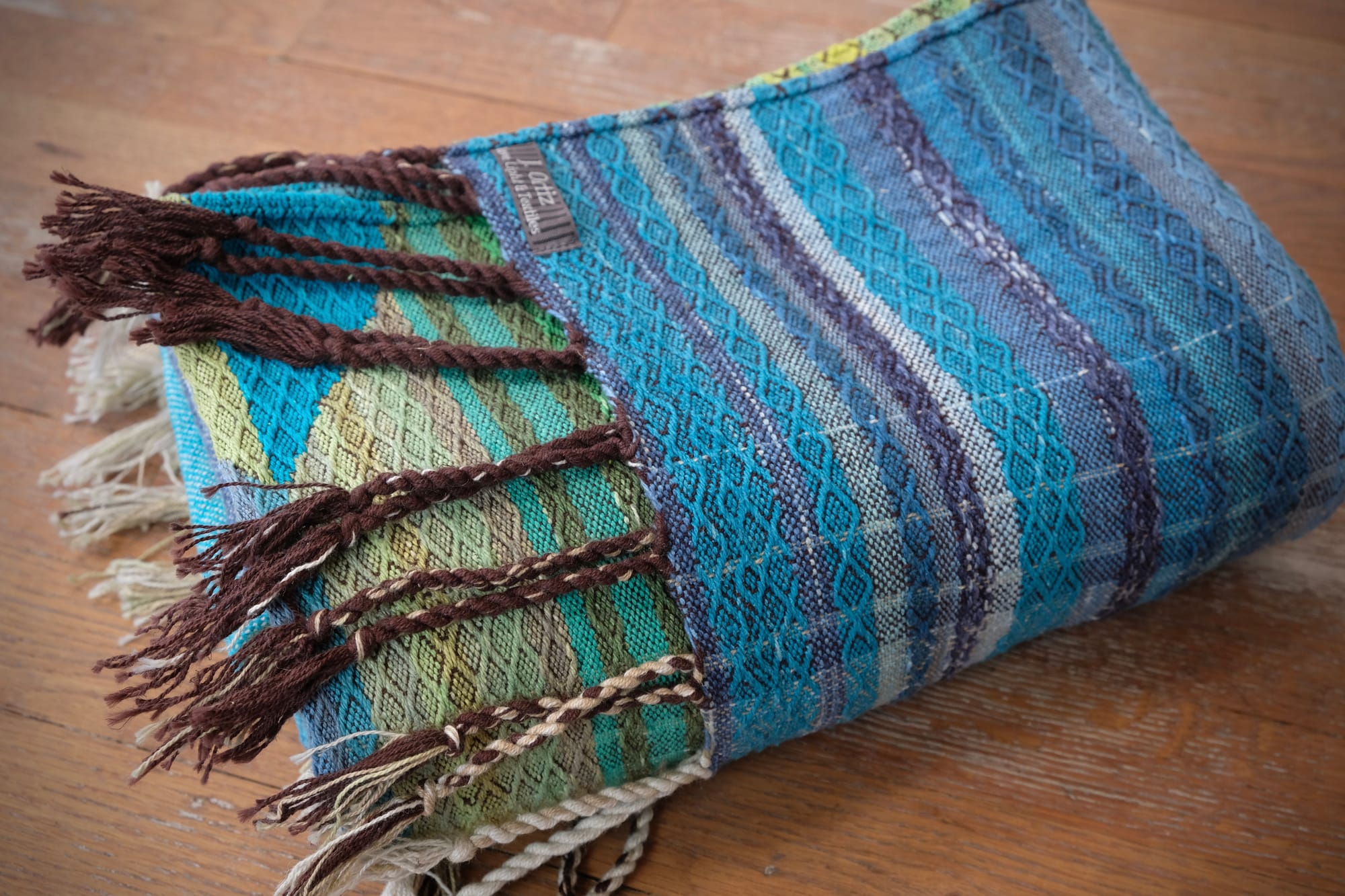 A folded bundle of handwoven fabric rests on an oak wood floor. The part of the fabric you can see is striped shades of blue and green with textured diamond pattern stripes