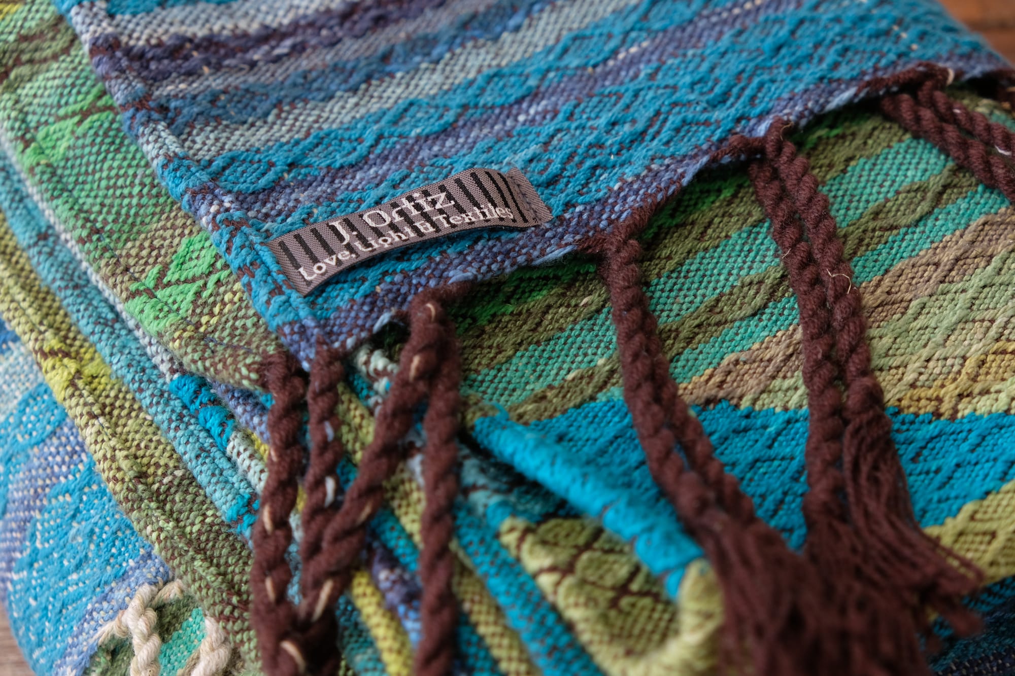 A folded bundle of handwoven fabric rests on an oak wood floor. The part of the fabric you can see is striped shades of blue and green with textured diamond pattern stripes