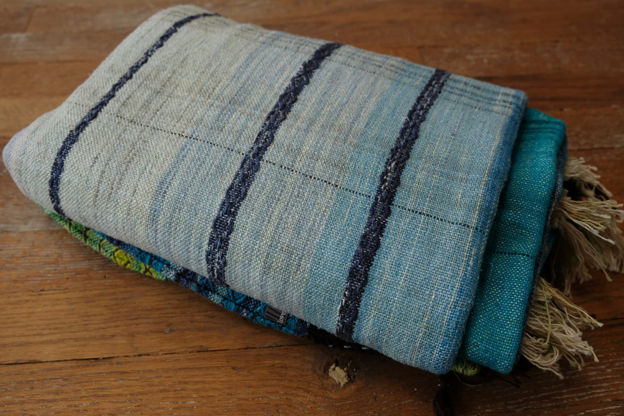 A folded bundle of handwoven fabric rests on an oak wood floor. The part of the fabric you can see is striped shades of blue with textured diamond pattern stripes