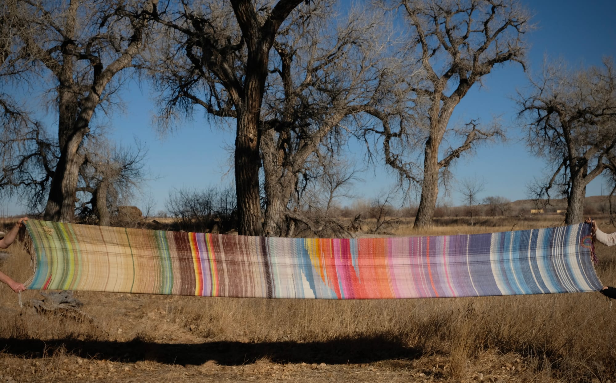 a long length of handwoven fabric in the colors of blue sky, sunrise and dry fields of barley, stretched against a golden landscape 