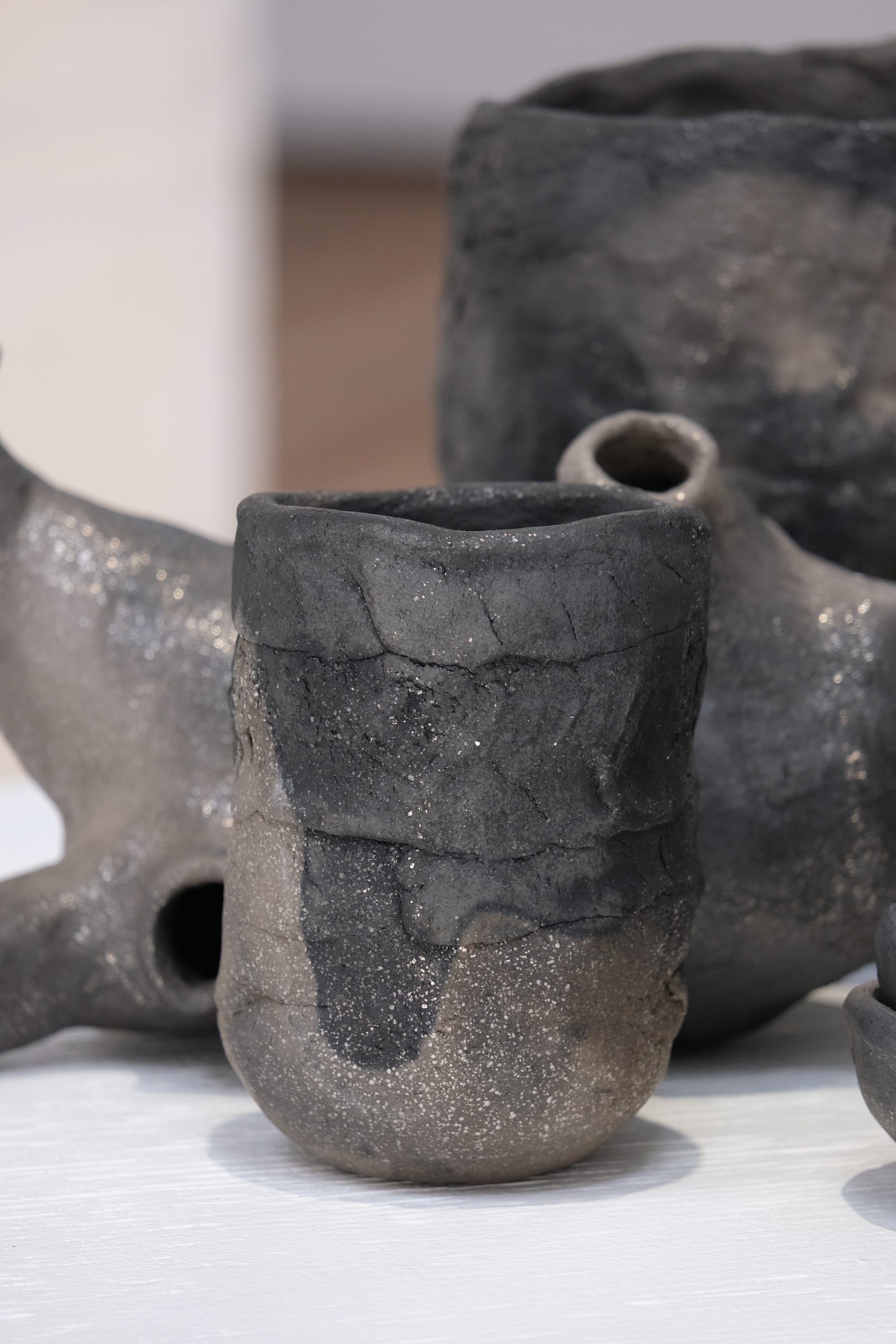 A close up view of a black and grey-brown sparkly clay vessel in an assemblage of other objets in an art installation on a white pedestal in a gallery 