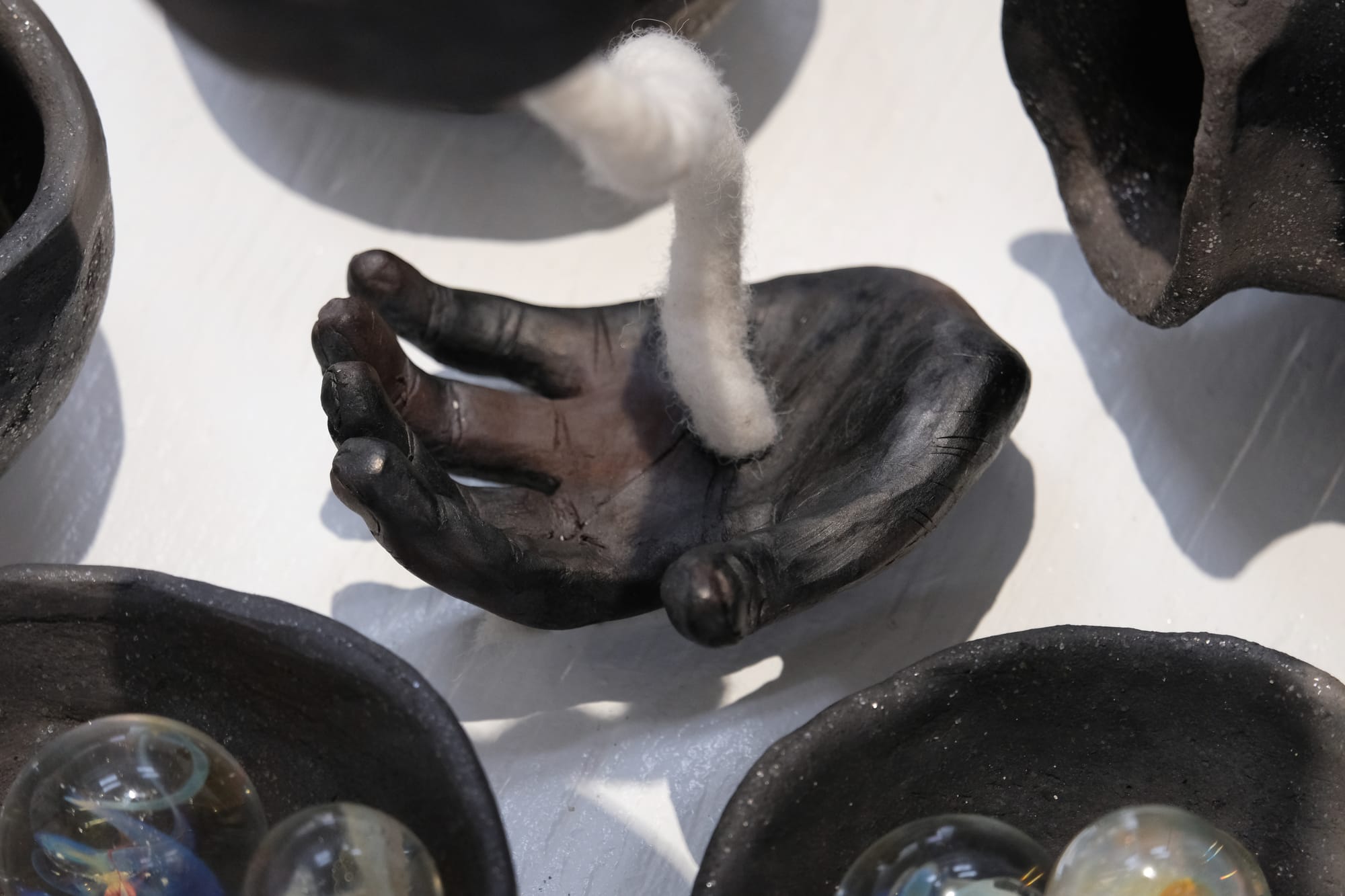 A close up view of a black and grey-brown sparkly clay vessel and hand sculpture in an assemblage of other objets in an art installation on a white pedestal in a gallery 