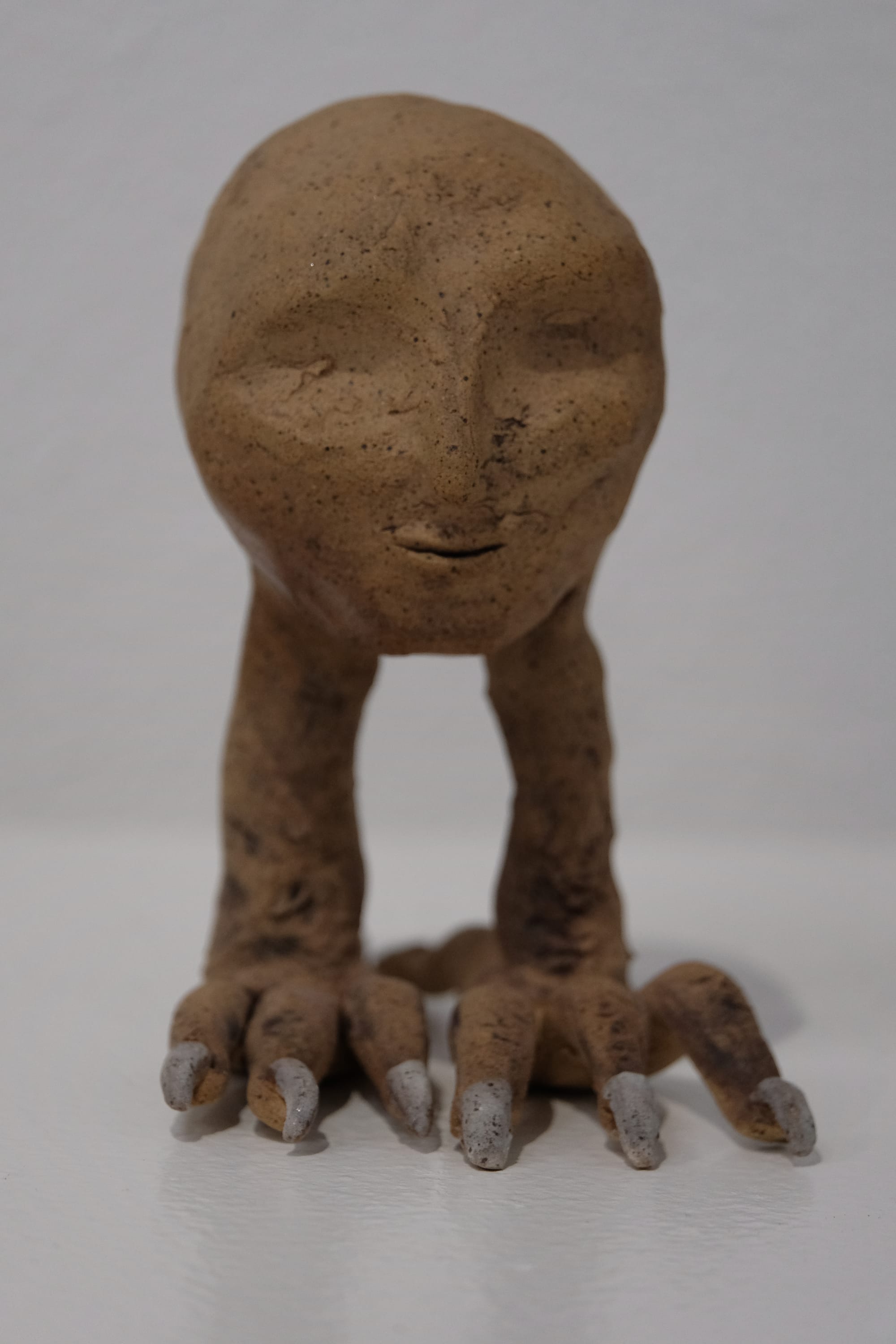 A small clay sculpture of a human head with raven legs. The clay is tan with dark speckles, the toenails of the creature are painted white