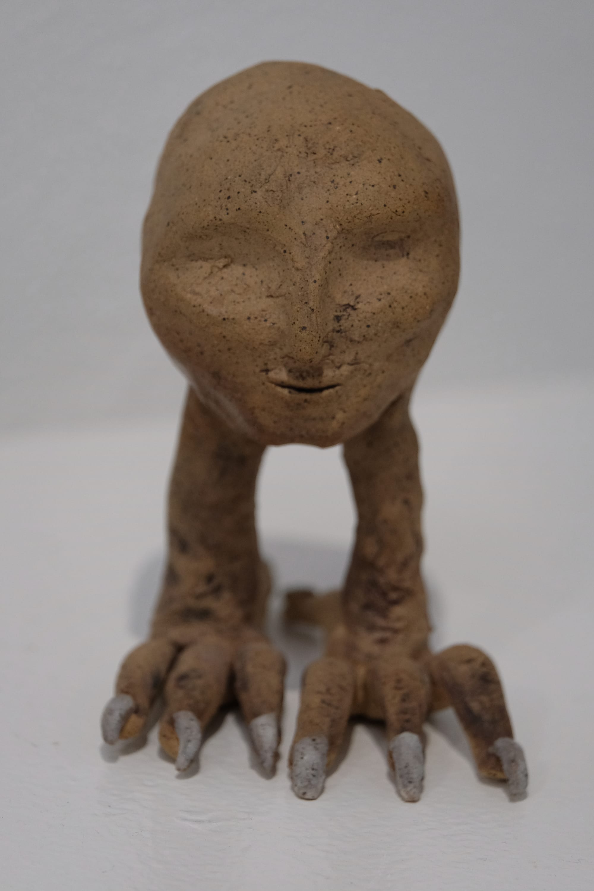 a small sculpture of a human head with bird legs stands on a white pedestal. it is made of tan clay with black speckles. the toenails are painted white 