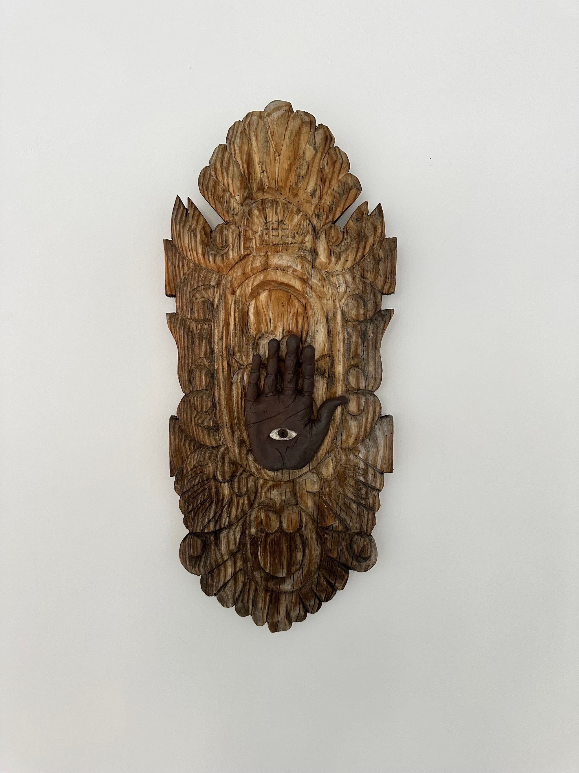 A sculpture on a white wall of a dark hand with an eye in the inside which is mounted on an intricately carved wooden tablet