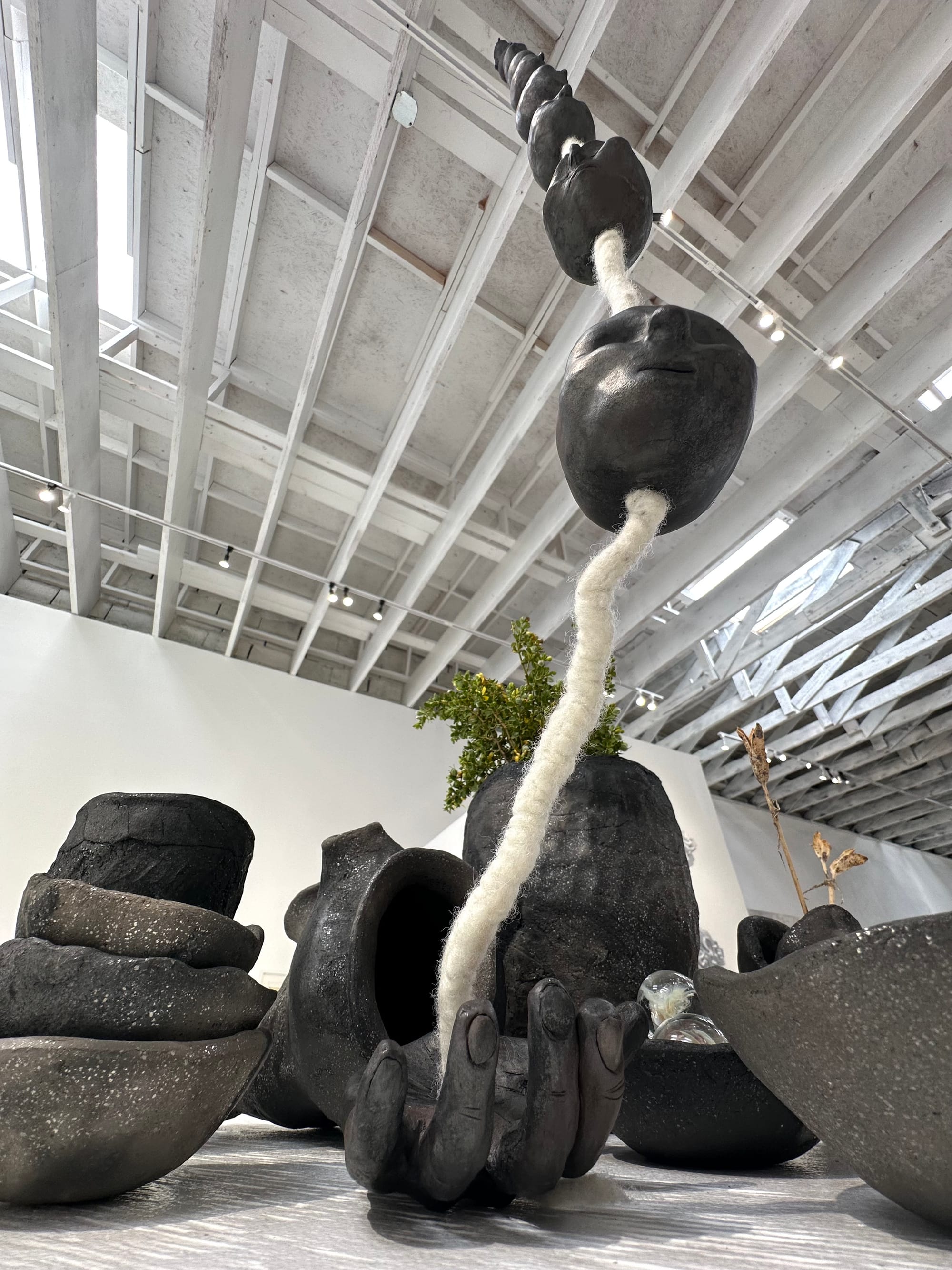 A close up view of a black and grey-brown sparkly clay vessel and hand sculpture in an assemblage of other objets in an art installation on a white pedestal in a gallery 