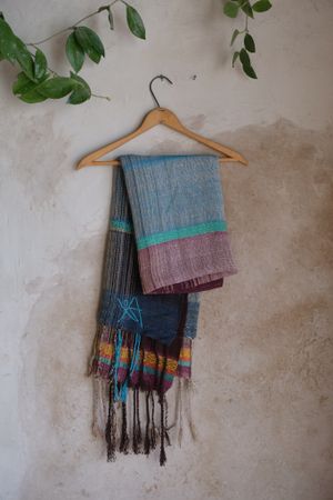 Handwoven maroon, blue, yellow and teal scarf on a hanger hanging on a tan wall.