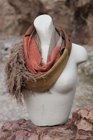 handwoven highly textured scarf that is mustard, brown, grey-blue and salmon colored, on a white mannequin sitting on a rock