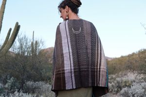 Woman wearing a handwoven black, pink, blue and lavender shawl with a crescent moon on it, standing in a desert landscape. 