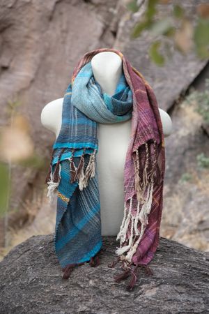 Handwoven maroon, blue, purple and yellow scarf on a white mannequin sitting on a boulder