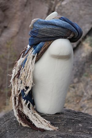 Blue, white and brown handwoven scarf on a white mannequin sitting on a boulder.