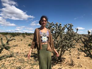 woman wearing handmade brown jacket and green skirt, standing in the desert with cactus