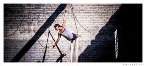 Woman in a grey shirt dong aerial acrobatics in front of a grey brick wall. 