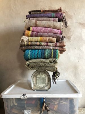 Stack of handwoven fabric in all colors, sitting on an old postal scale, on a clear tote. 
